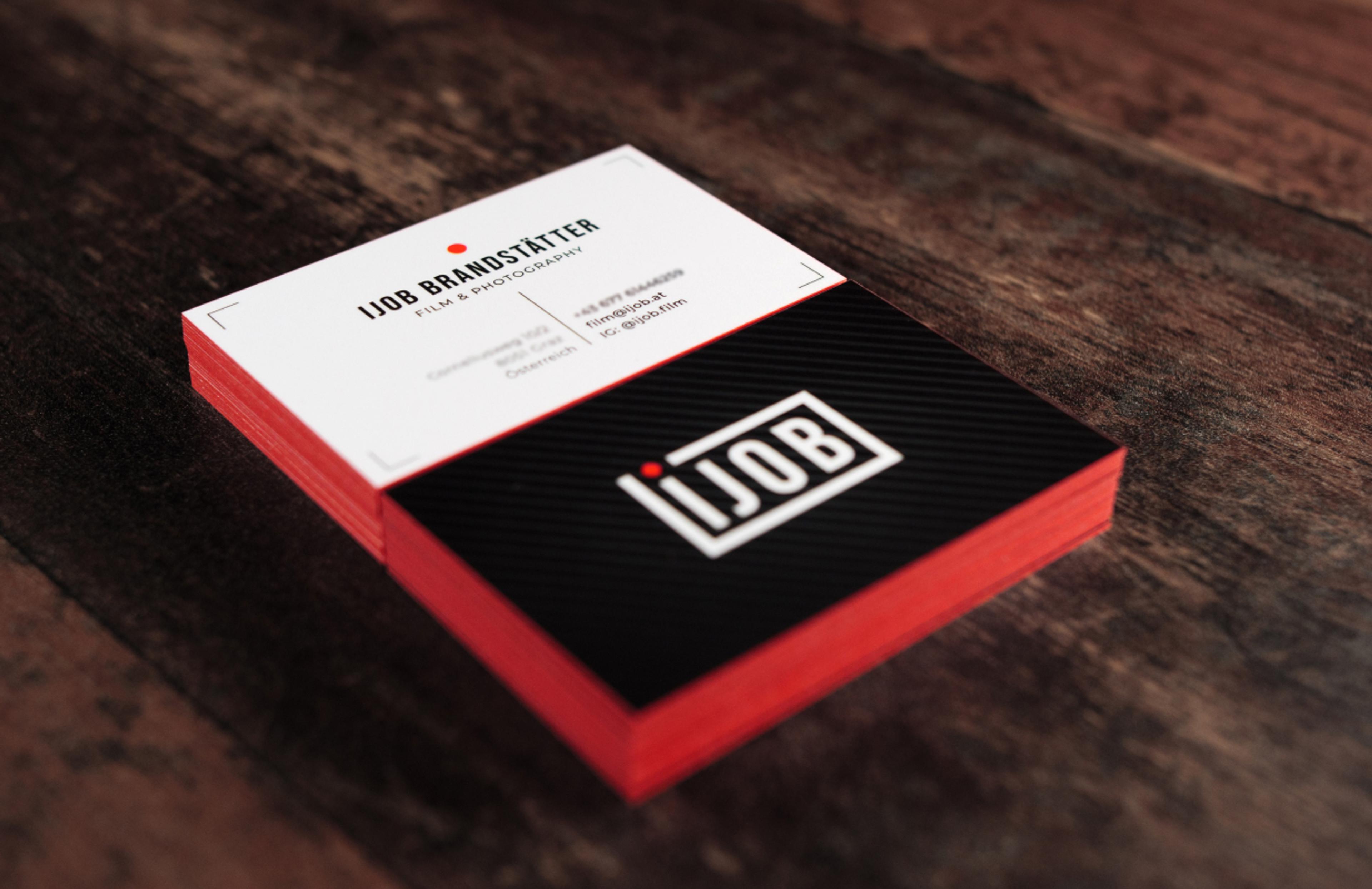Mehrere Visitenkarten in Schwarz-Weiß mit roten Akzenten. Die Karten enthalten das Ijob-Logo, den Namen „Ijob Brandstätter“ und Kontaktdaten, platziert auf einer strukturierten, schwarz gestreiften Oberfläche.