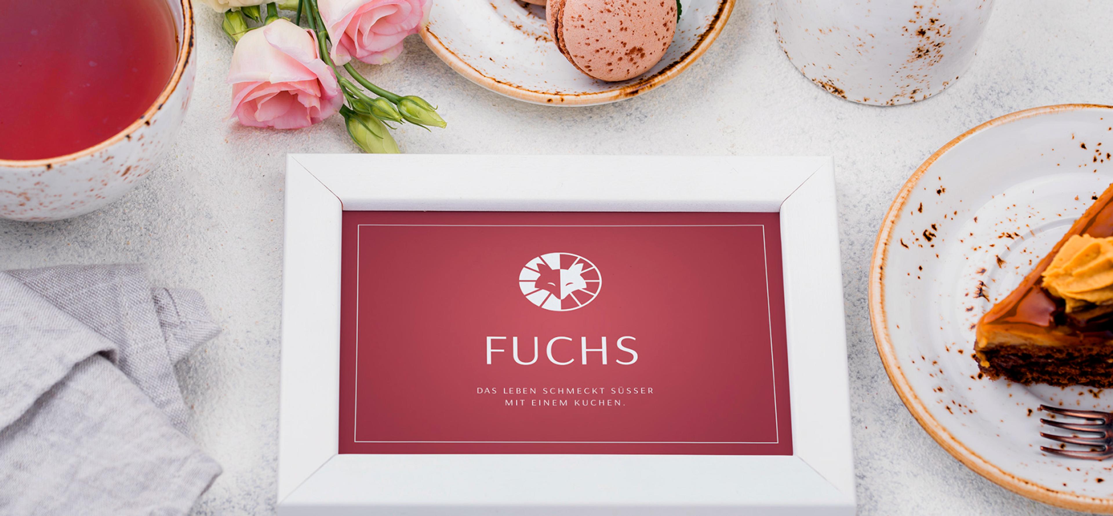 Ein weißer Rahmen mit dem „Fuchs“-Logo und dem Slogan „Das Leben schmeckt süßer mit einem Kuchen“, umgeben von einer Tasse Tee, rosa Blumen und einem Teller mit Macarons.