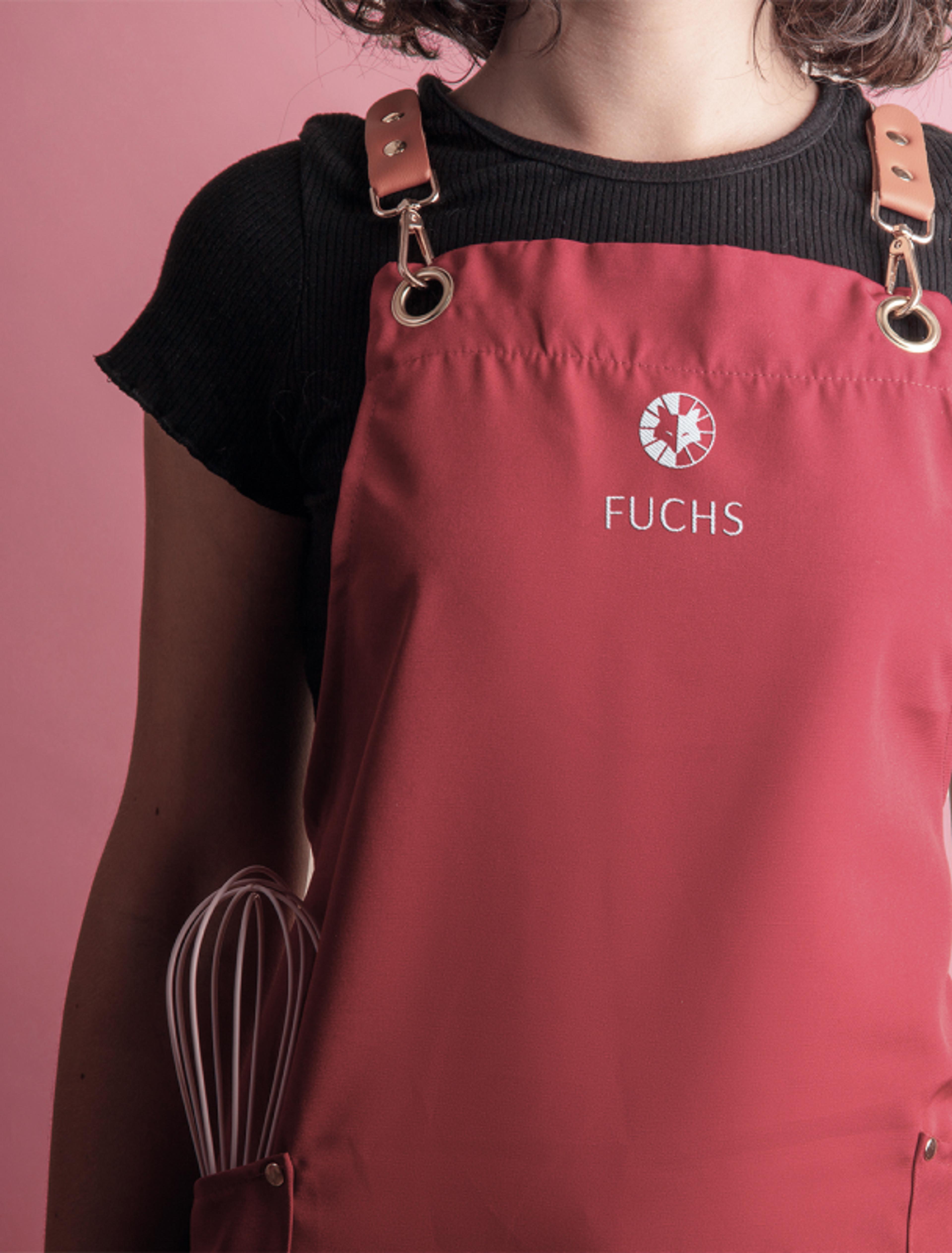 Eine Frau trägt eine dunkelrote Schürze mit hochwertiger Stickerei des „Fuchs“-Logos. Die Lederriemen und Metallösen verleihen der Schürze einen edlen Look.