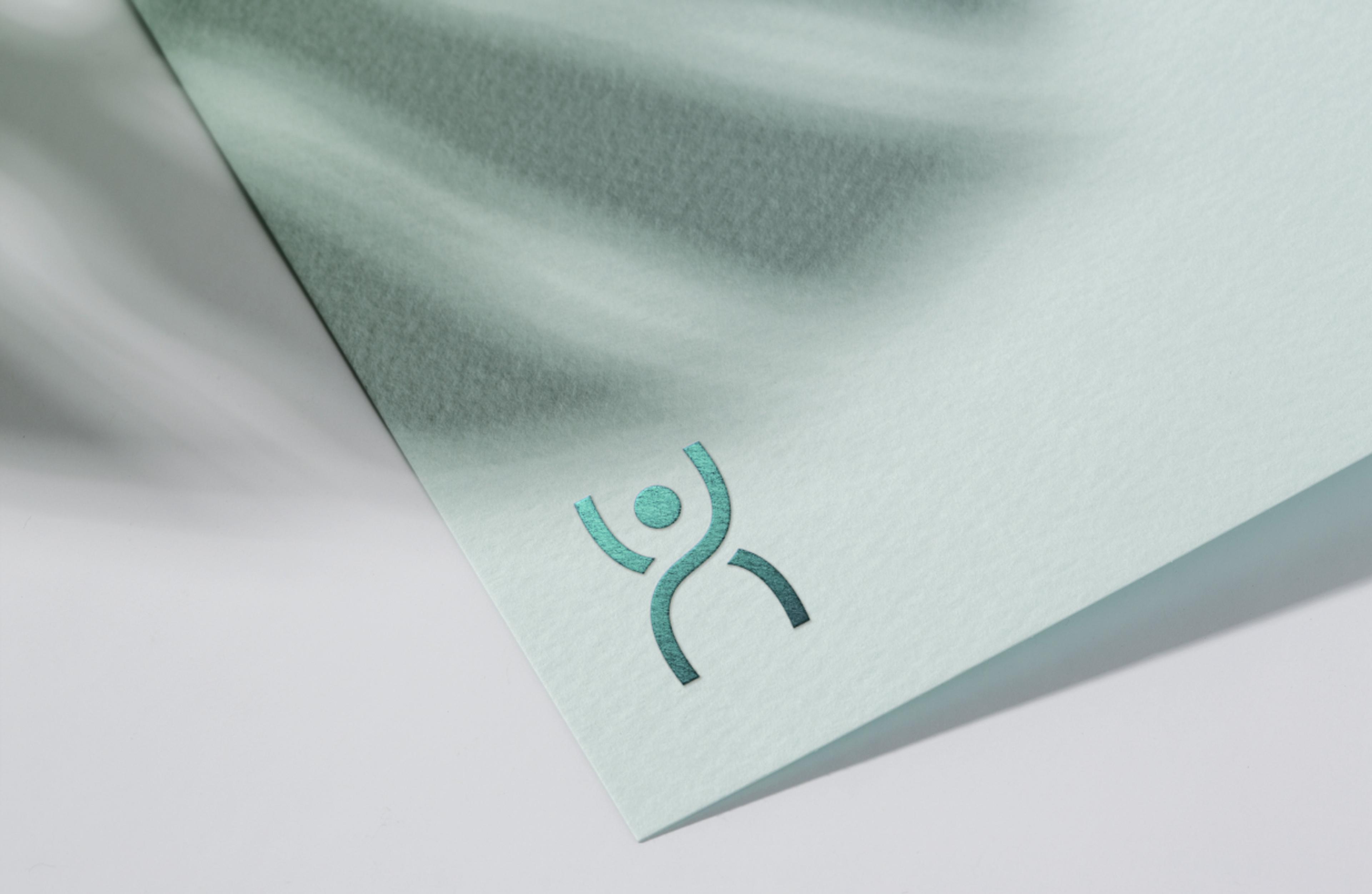 Hochwertig geprägtes Logo auf strukturiertem Papier mit schimmerndem Finish. Diese edle Verarbeitung unterstreicht den Premium-Charakter der Marke. Die grafische Formsprache des Logos – ein stilisierter Mensch in Bewegung – symbolisiert Therapie, Dynamik und Fortschritt.