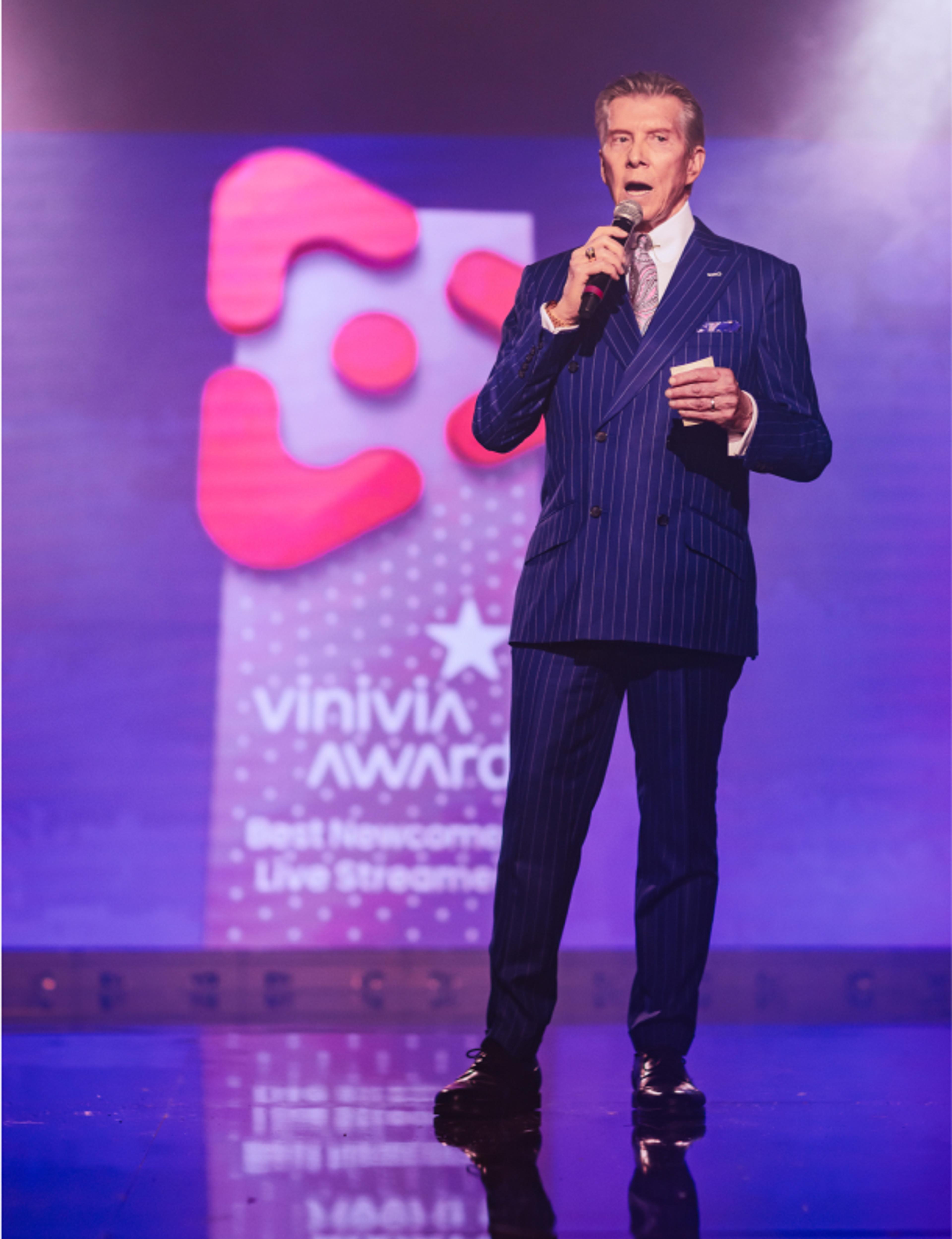 Foto: vinivia, bfa, jorgemezaphotos Moderator auf der Vinivia Awards Bühne: Live-Auftritt vor großem Logo-Screen in violettem Event-Design – Unerwartet Design