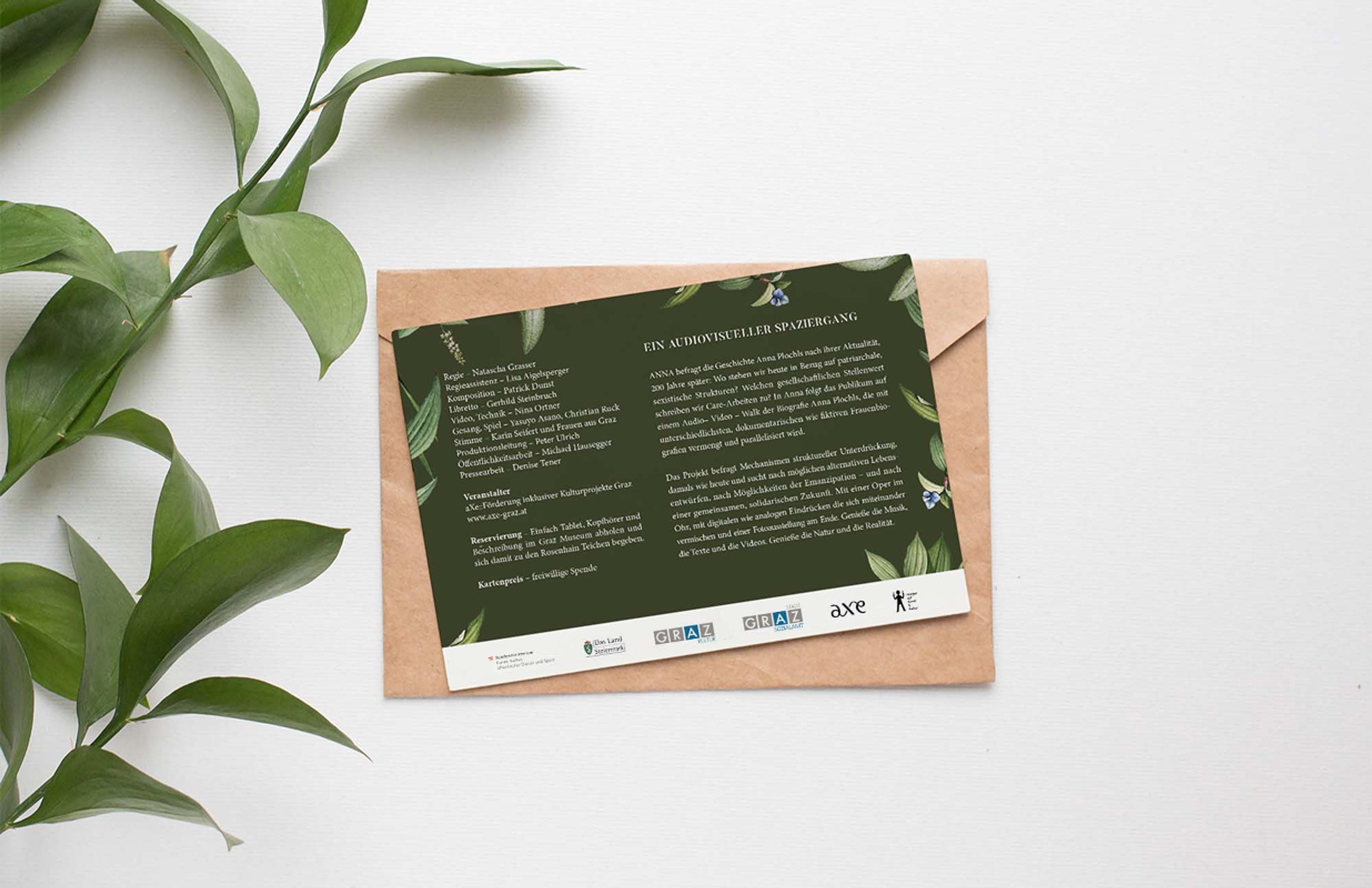 Grüner Flyer auf Umschlag mit Pflanzen – Ein dunkelgrüner Flyer mit floralen Elementen liegt auf einem braunen Umschlag, umgeben von grünen Blättern.