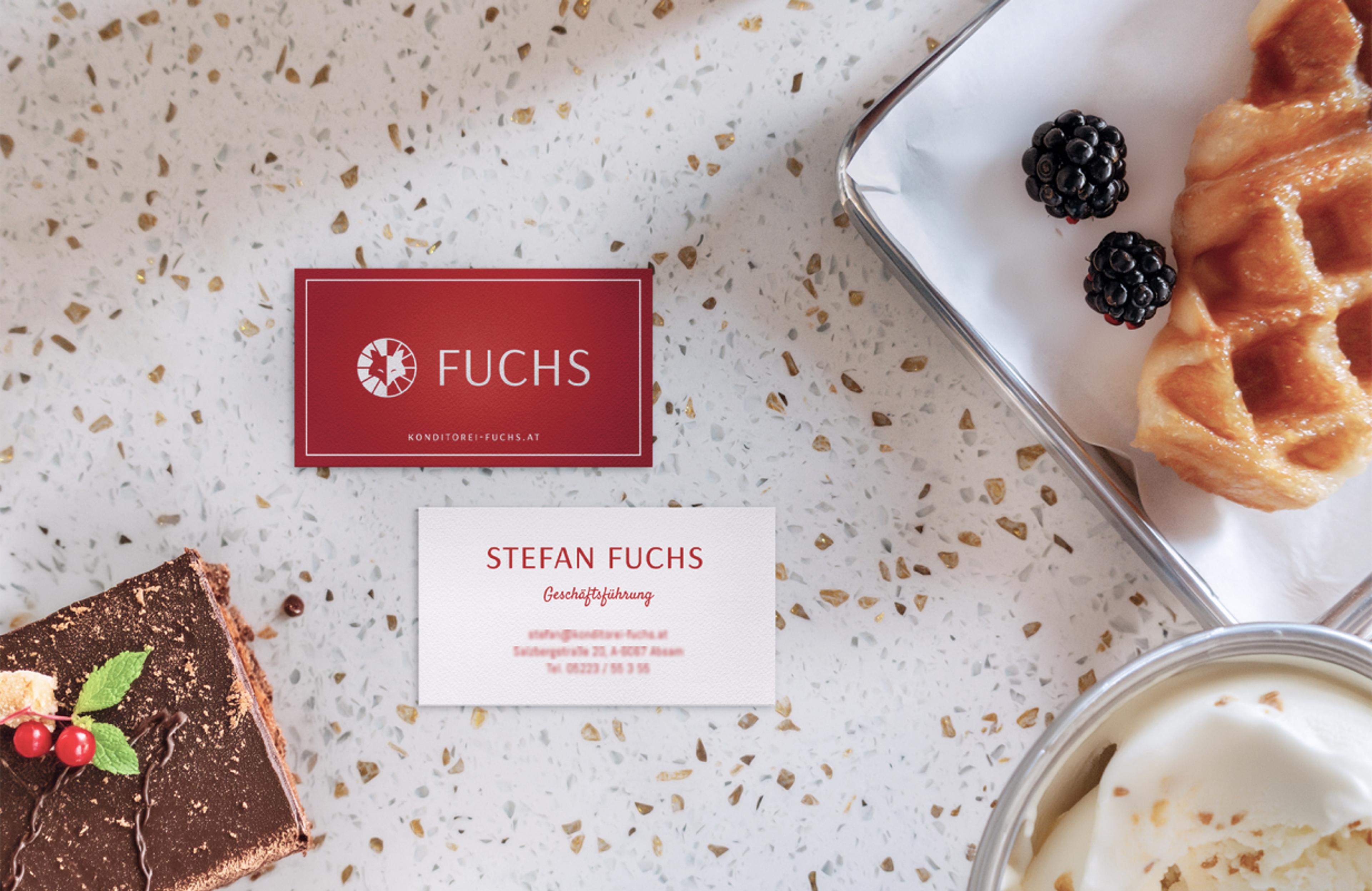 Zwei Visitenkarten, eine mit dem roten „Fuchs“-Logo und eine mit den Geschäftsinformationen von Stefan Fuchs. Daneben liegen eine Schokoladentorte, eine Waffel mit Beeren und ein Glas Dessert.