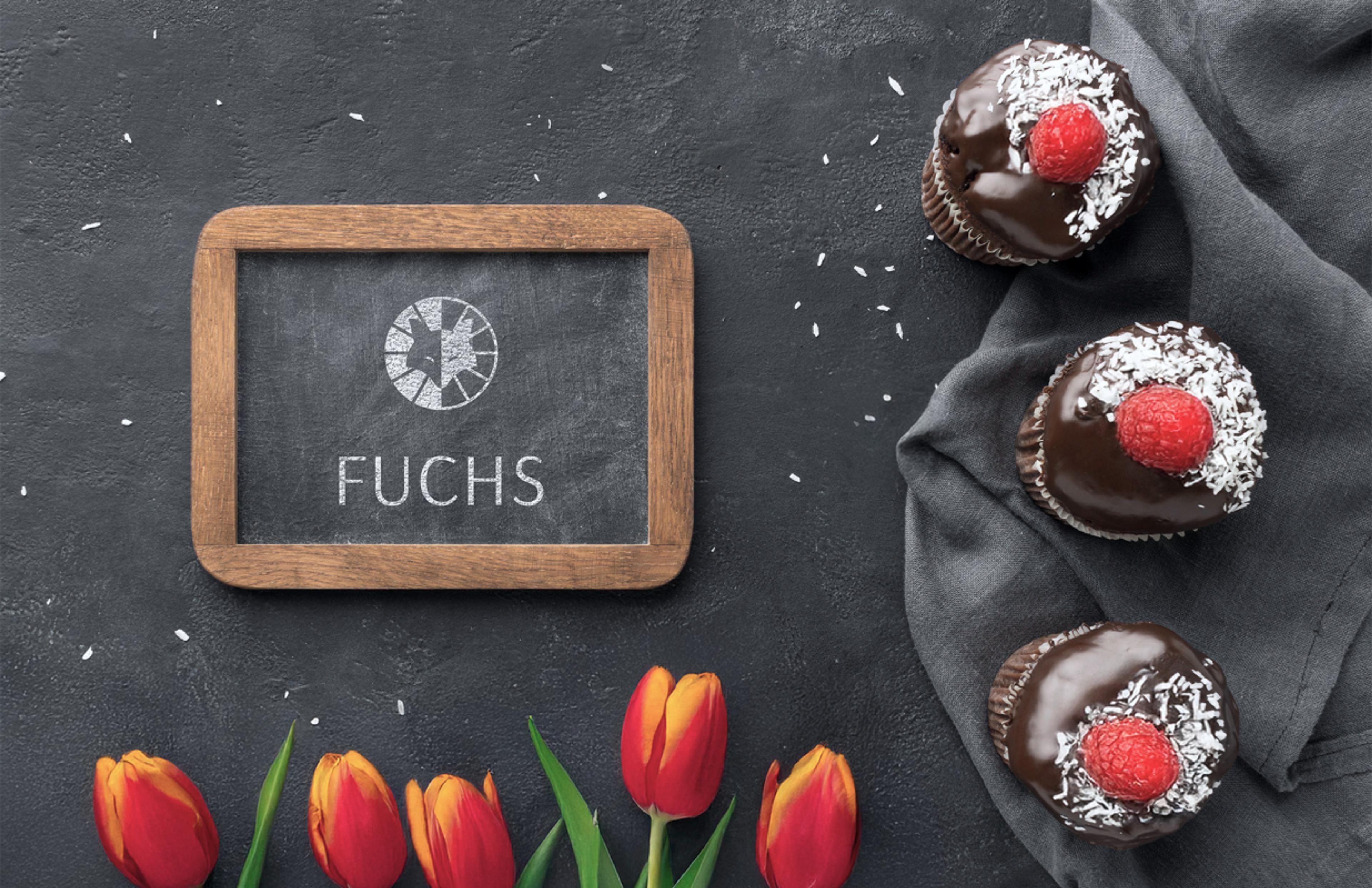 Eine kleine Holztafel mit dem „Fuchs“-Logo in Kreideoptik, umgeben von drei mit Schokolade glasierten Muffins mit Kokosraspeln und Himbeeren sowie frischen Tulpen.