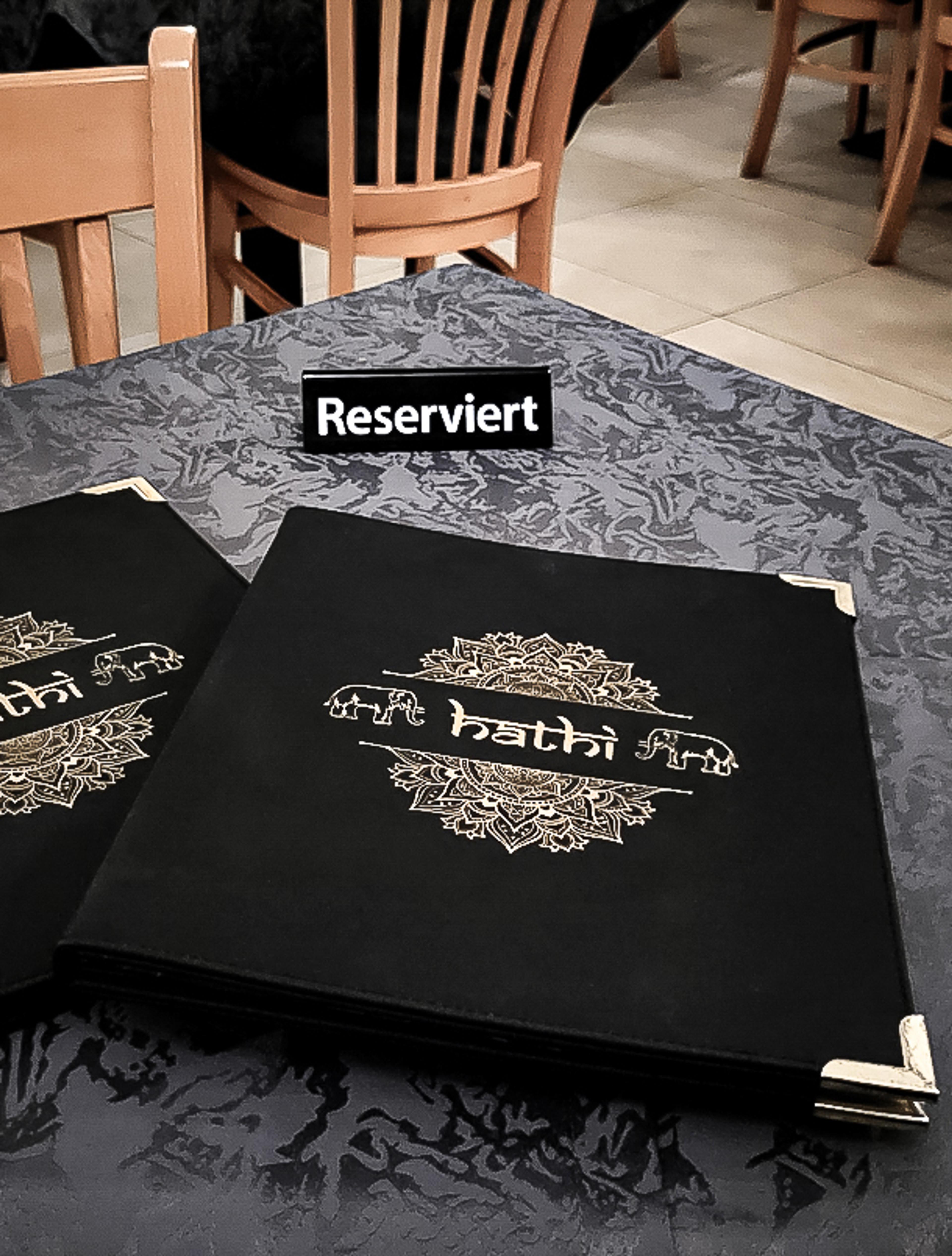Exklusiv reservierter Tisch im Hathi indischen Restaurant mit hochwertiger Speisekarte in Schwarz und Gold. Die edle Tischgestaltung kombiniert dunkle Stoffe mit traditionellen indischen Mustern und feinen Details. Perfektes Gastronomie-Branding für luxuriöse Restaurants, umgesetzt von Unerwartet Branding und Design OG aus Graz, Österreich.