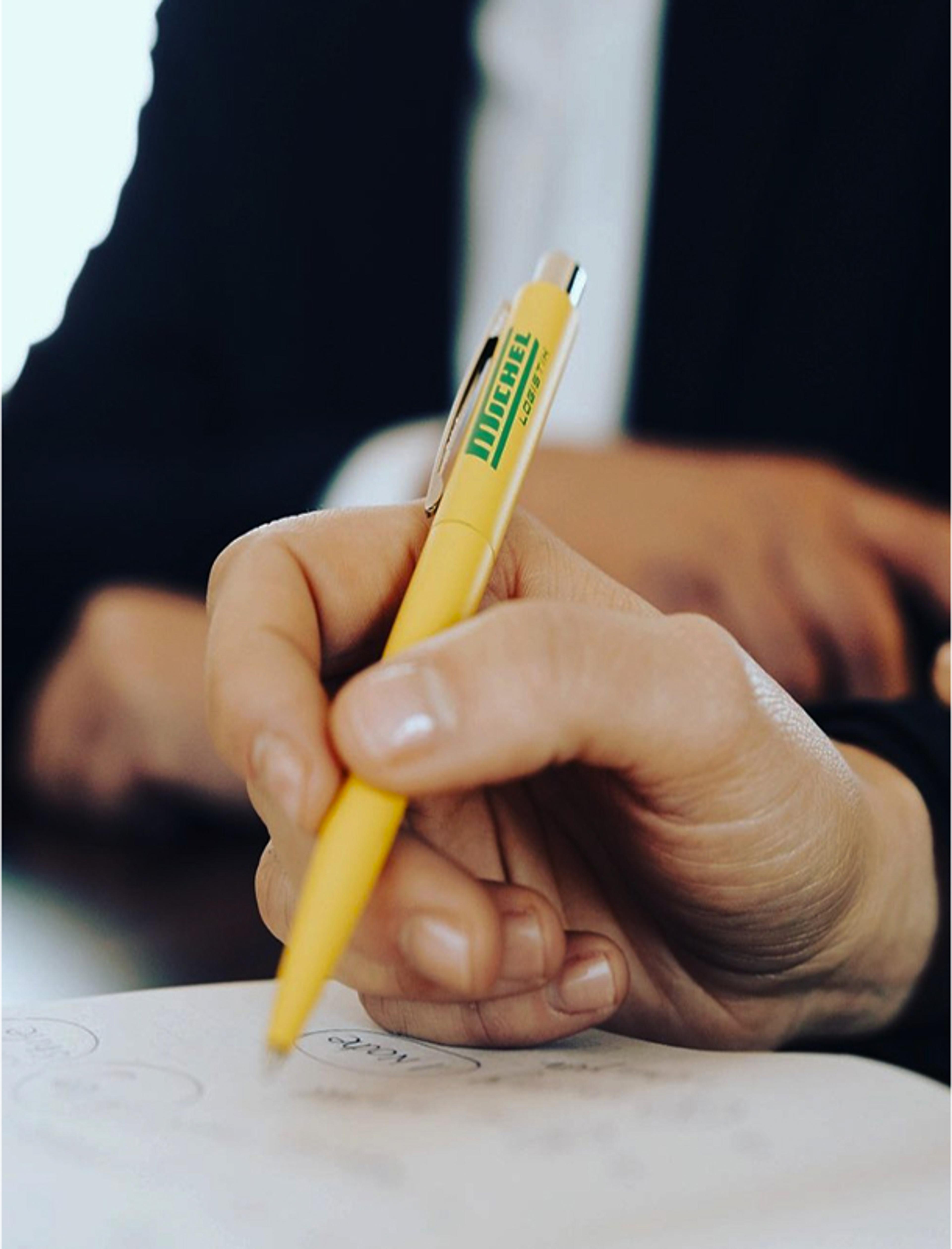 Corporate Design für Michel Logistik – Kugelschreiber Branding Nahaufnahme eines gelben Michel Logistik Kugelschreibers in der Hand. Modernes Markenbranding und Corporate Design für die Transport- und Logistikbranche, entwickelt von Unerwartet Design.