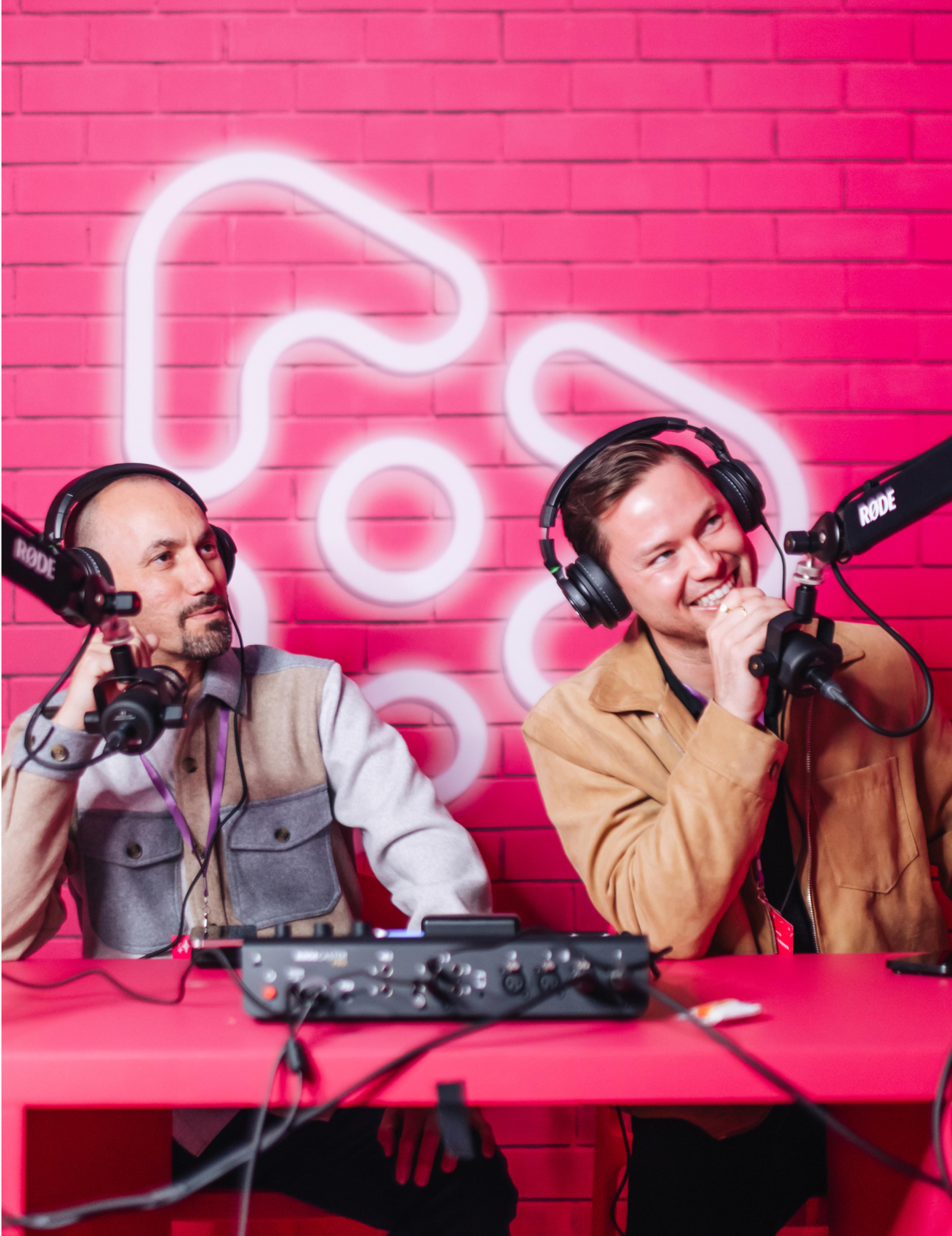 Foto: vinivia, bfa, jorgemezaphotos Zwei Podcaster in der Vinivia Streaming-Lounge vor pinker Backsteinwand mit beleuchtetem Logo – Unerwartet Design
