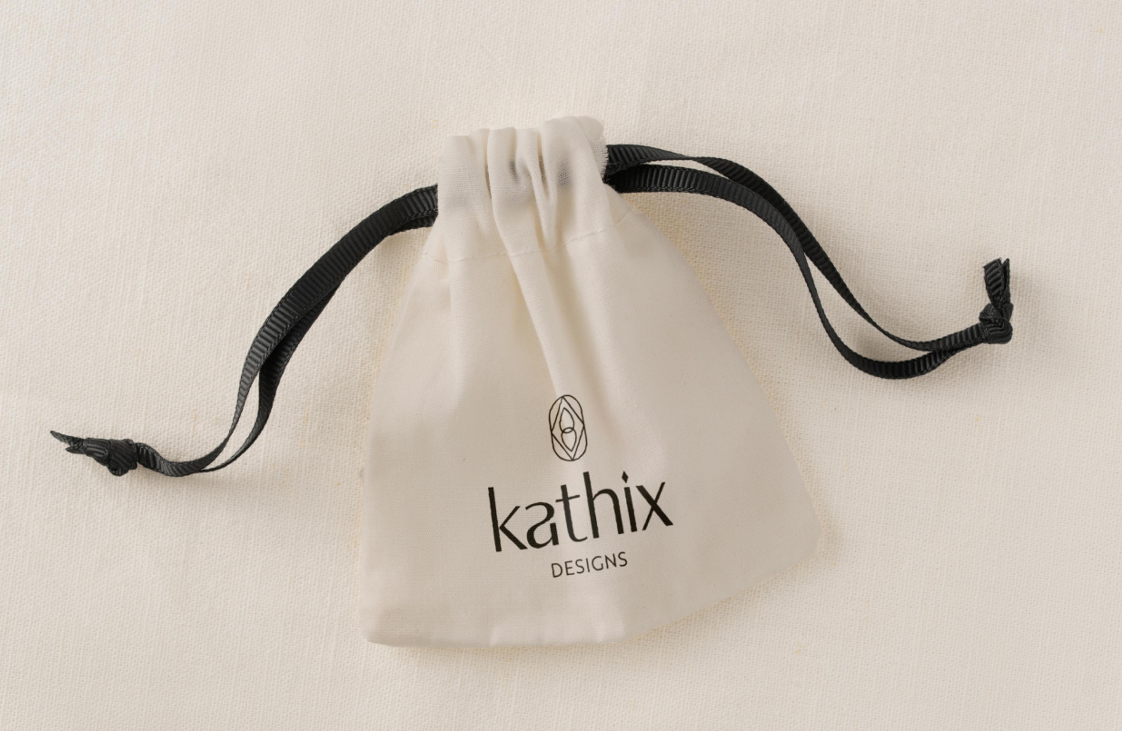 Eleganter Stoffbeutel mit Kathix Designs Logo – Ein hochwertiger, naturfarbener Stoffbeutel mit schwarzem Logo-Print und schwarzen Zugbändern, passend für luxuriöse Verpackungen oder Accessoires.