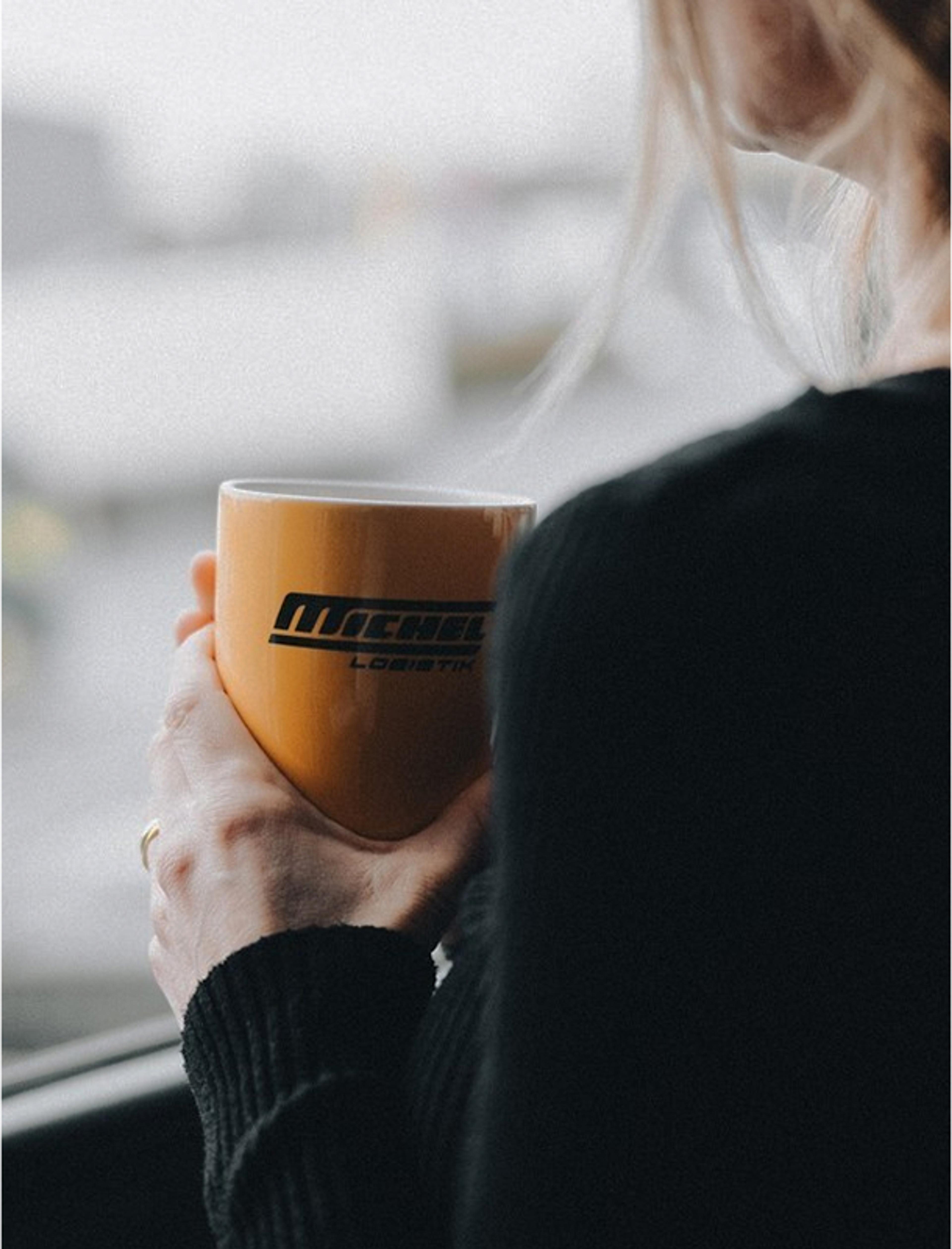 Eine Frau hält eine gelbe Michel Logistik Kaffeetasse. Logo-Design und Markenstrategie, die Vertrauen und Identität in der Transportbranche stärken.
