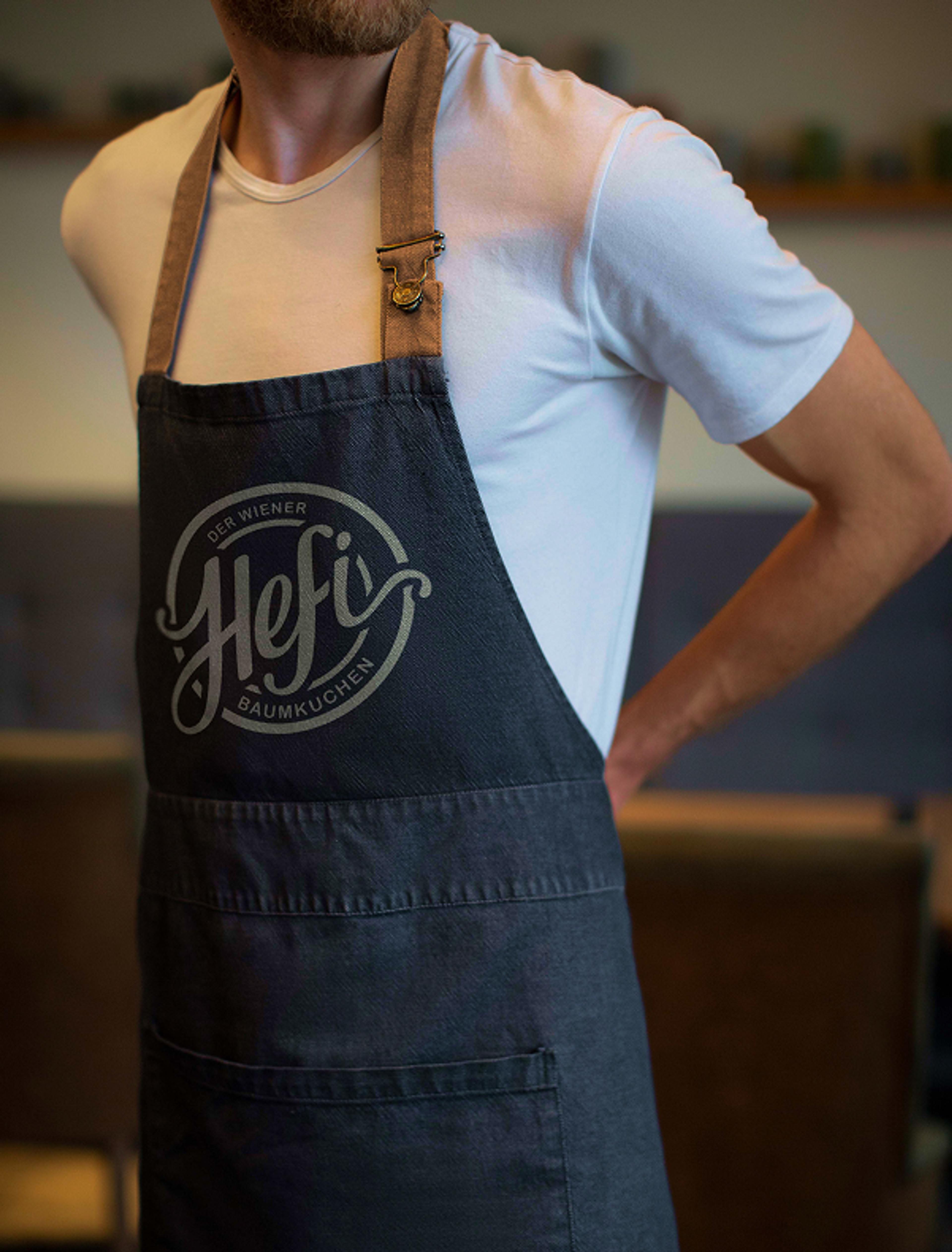 Ein Mitarbeiter trägt eine dunkelgraue Schürze mit dem Hefi-Logo in einem Café.