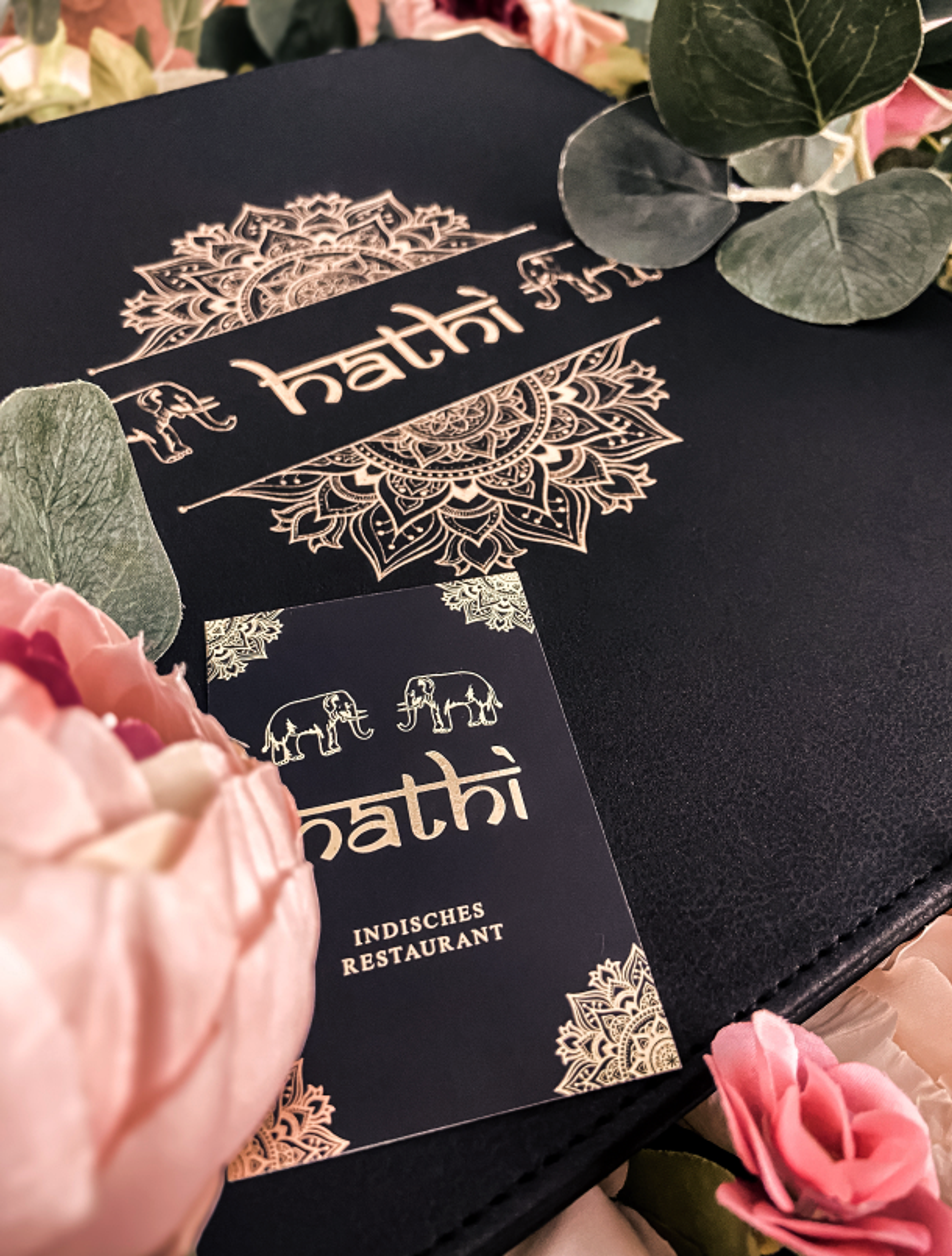 Stilvolle Speisekarte und Visitenkarte des Hathi indischen Restaurants, kunstvoll inszeniert mit Blumen und Blättern. Edles Logo mit goldener Prägung, verziert mit Mandala-Mustern und Elefantenmotiven. Hochwertiges Corporate Design für gehobene indische Gastronomie, entwickelt von Unerwartet Branding und Design OG aus Graz, Österreich.