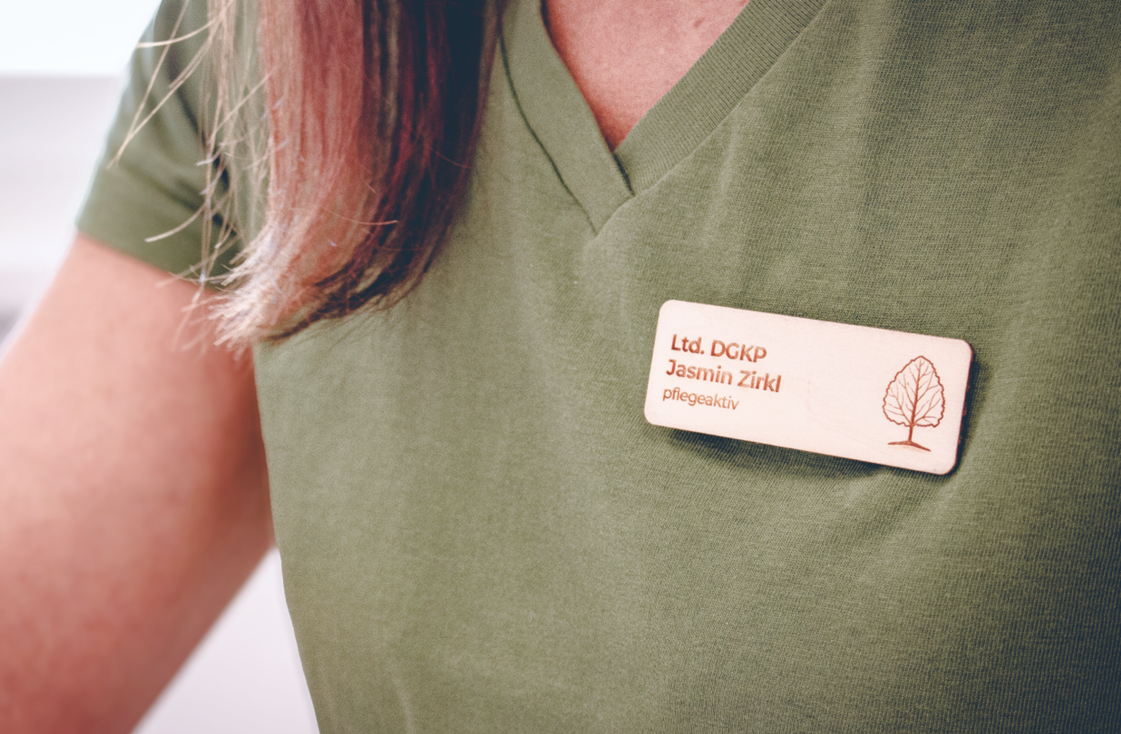 Logo & Grundkonzept BUCHER BRAND MARKETING e.U. Detailaufnahme eines gravierten Namensschilds aus Holz (Jasmin Zirkl) mit dem Logo des Lindenparks, getragen von einer Mitarbeiterin in grüner Uniform. Harmonisches Branding durch Unerwartet Design.