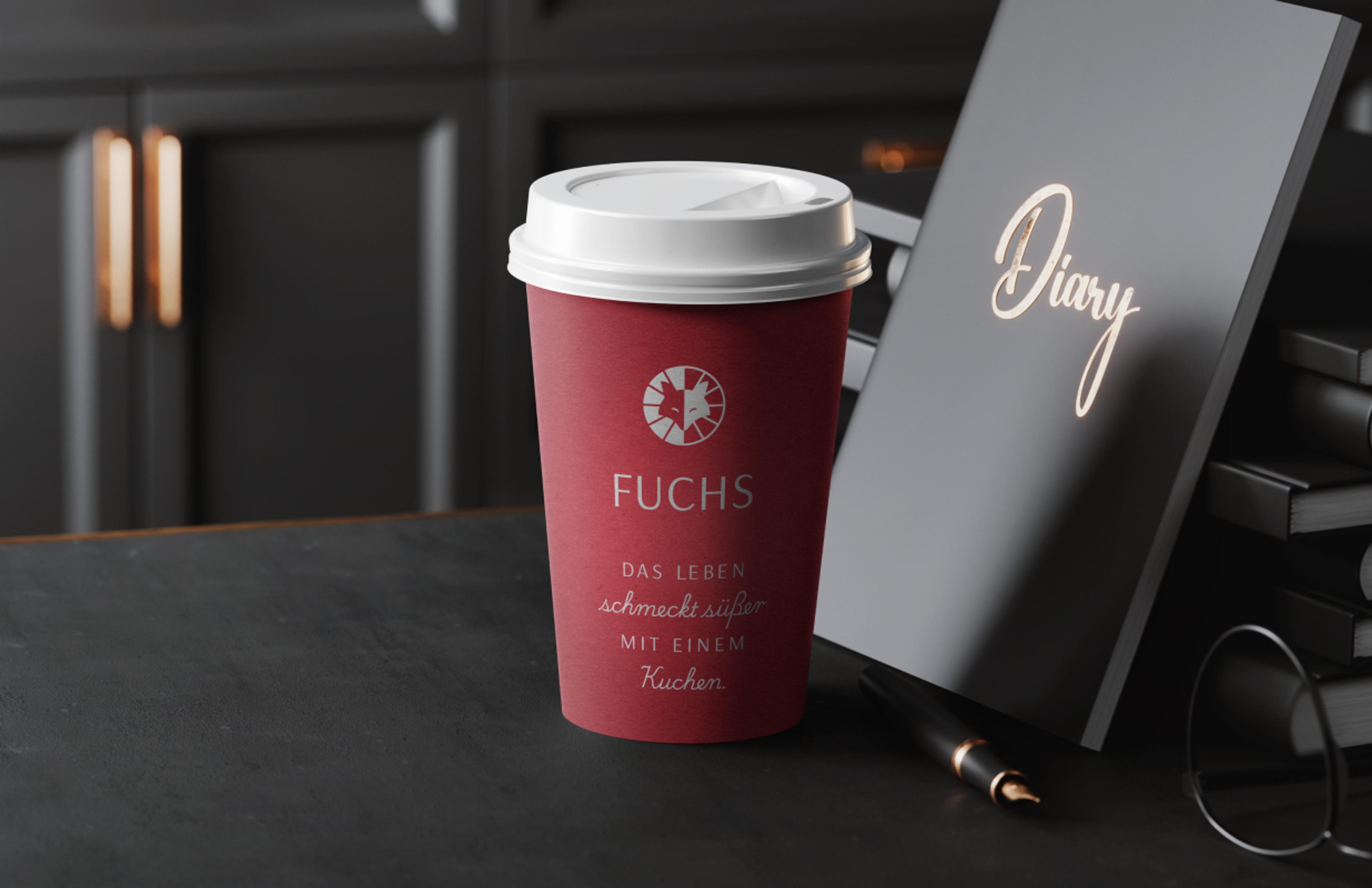 Ein dunkelroter Pappbecher mit weißem Deckel, verziert mit dem „Fuchs“-Logo und dem Slogan in eleganter Schrift. Daneben liegt ein Notizbuch mit „Diary“-Schriftzug.