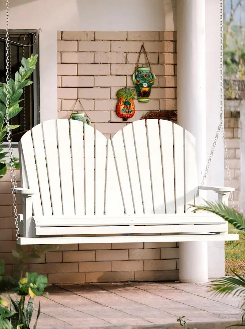 White double Adirondack porch swing with slatted back hanging from chains against brick wall.