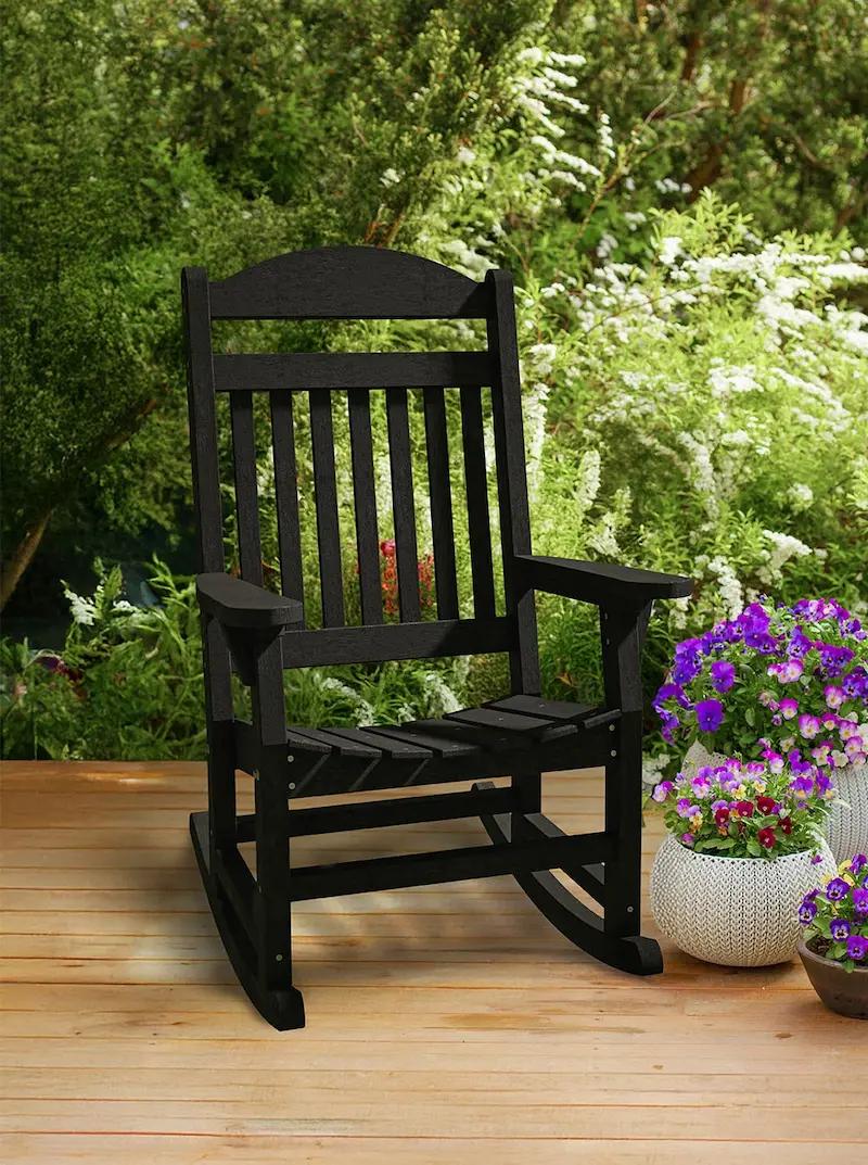 Black outdoor rocking chair with vertical slat backrest on a wooden deck surrounded by flowers.