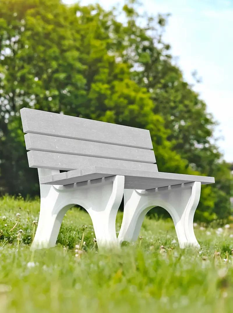 Gray and white outdoor bench with slatted backrest placed on grass with trees in background.