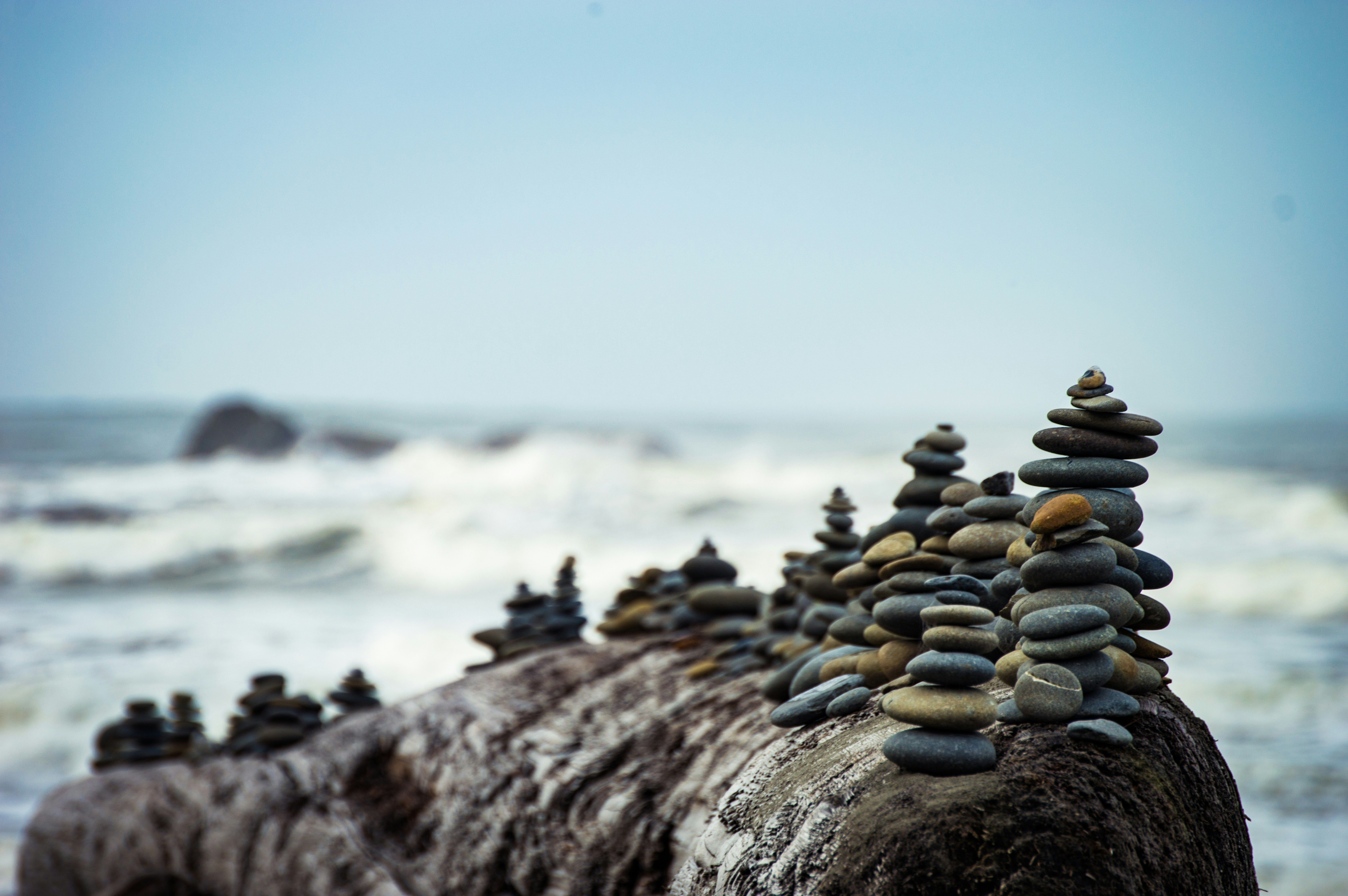 Stones stacked in contemplative towers on top a large boulder, white waves crashing in a background. Illustrating the need for calm strategic patience in the face of market chaos 