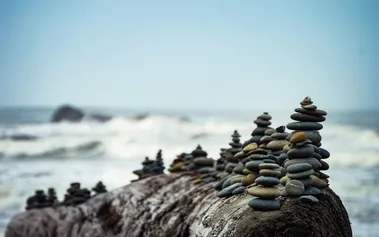 Stones stacked in contemplative towers on top a large boulder, white waves crashing in a background. Illustrating the need for calm strategic patience in the face of market chaos