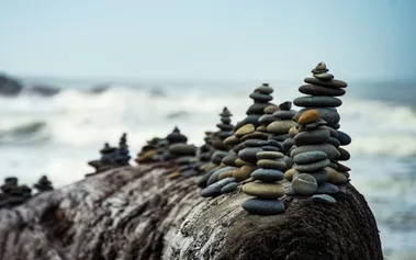 Stones stacked in contemplative towers on top a large boulder, white waves crashing in a background. Illustrating the need for calm strategic patience in the face of market chaos