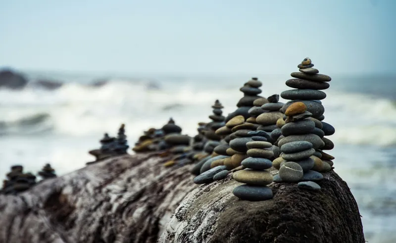 Stones stacked in contemplative towers on top a large boulder, white waves crashing in a background. Illustrating the need for calm strategic patience in the face of market chaos