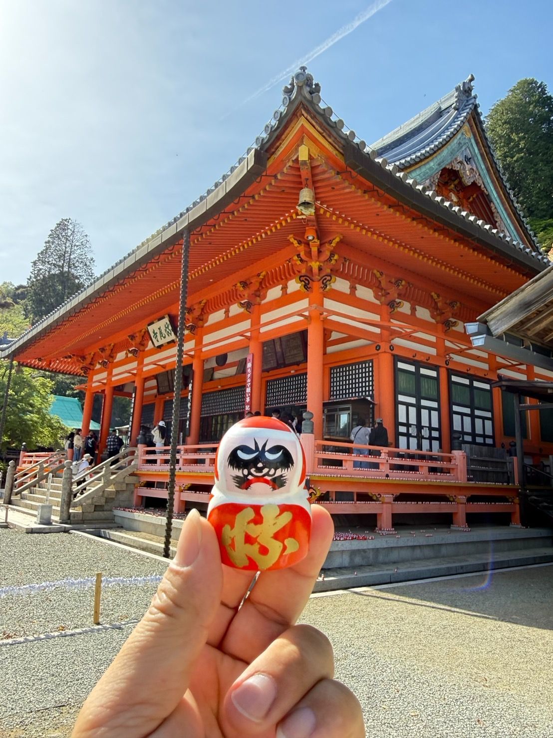 本堂旁的「迷你達摩見本堂」經典角度——手拿小達摩,背景是紅色大寺