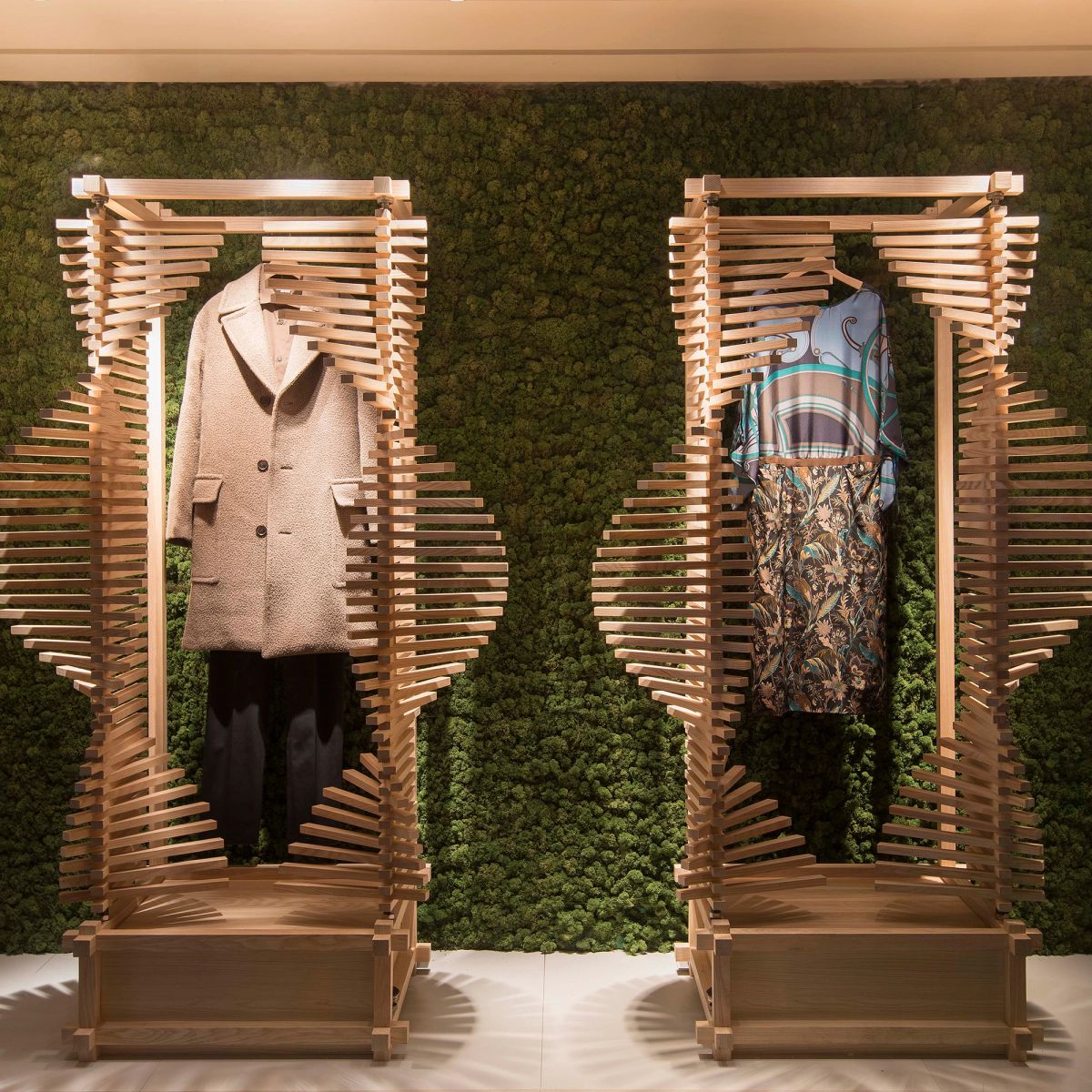 Two spiral wooden racks display a beige coat and a patterned dress against a green moss wall.