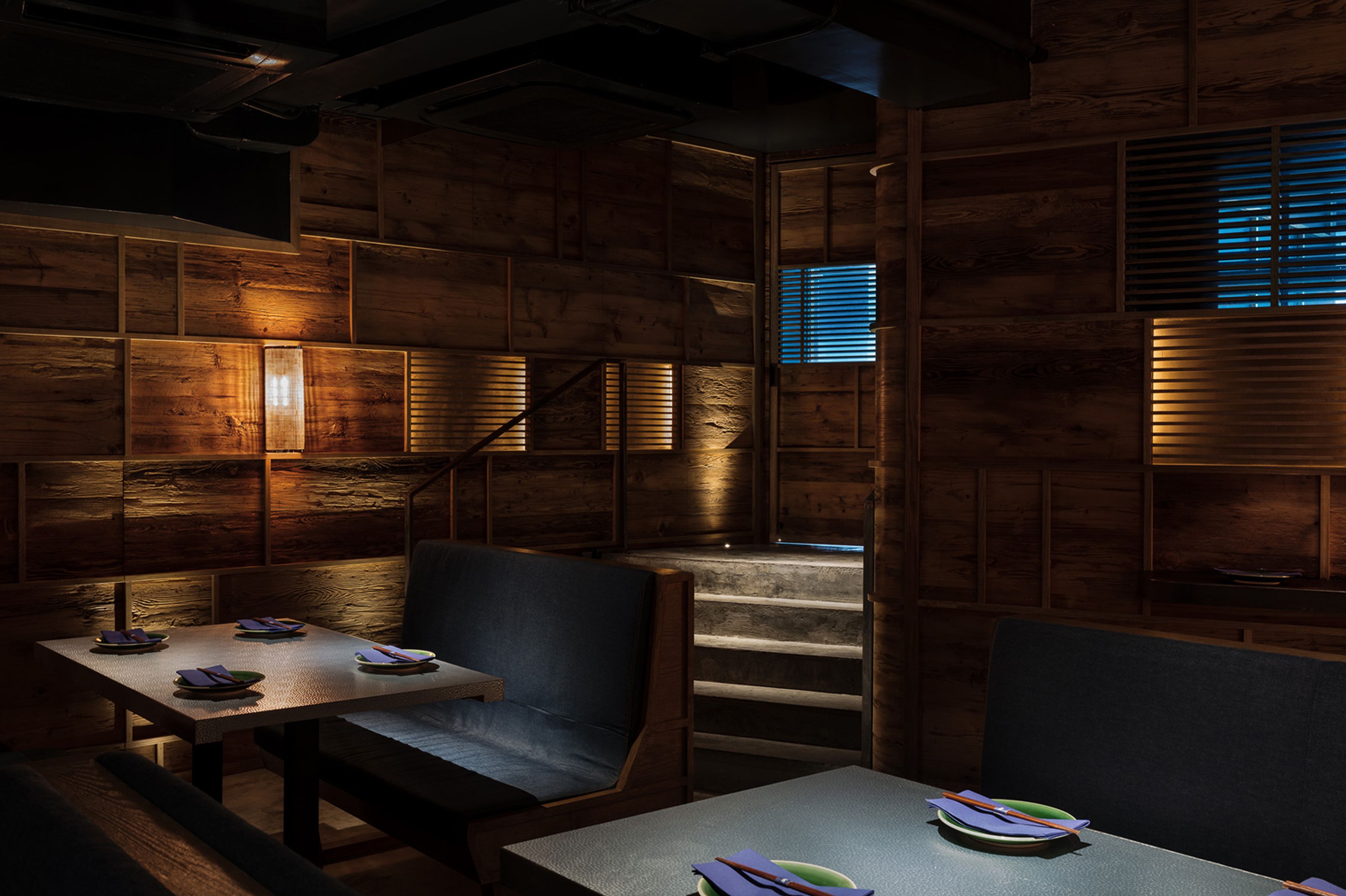 Dimly lit restaurant with wood-paneled walls, booths, and tables set with plates, leading to a staircase.