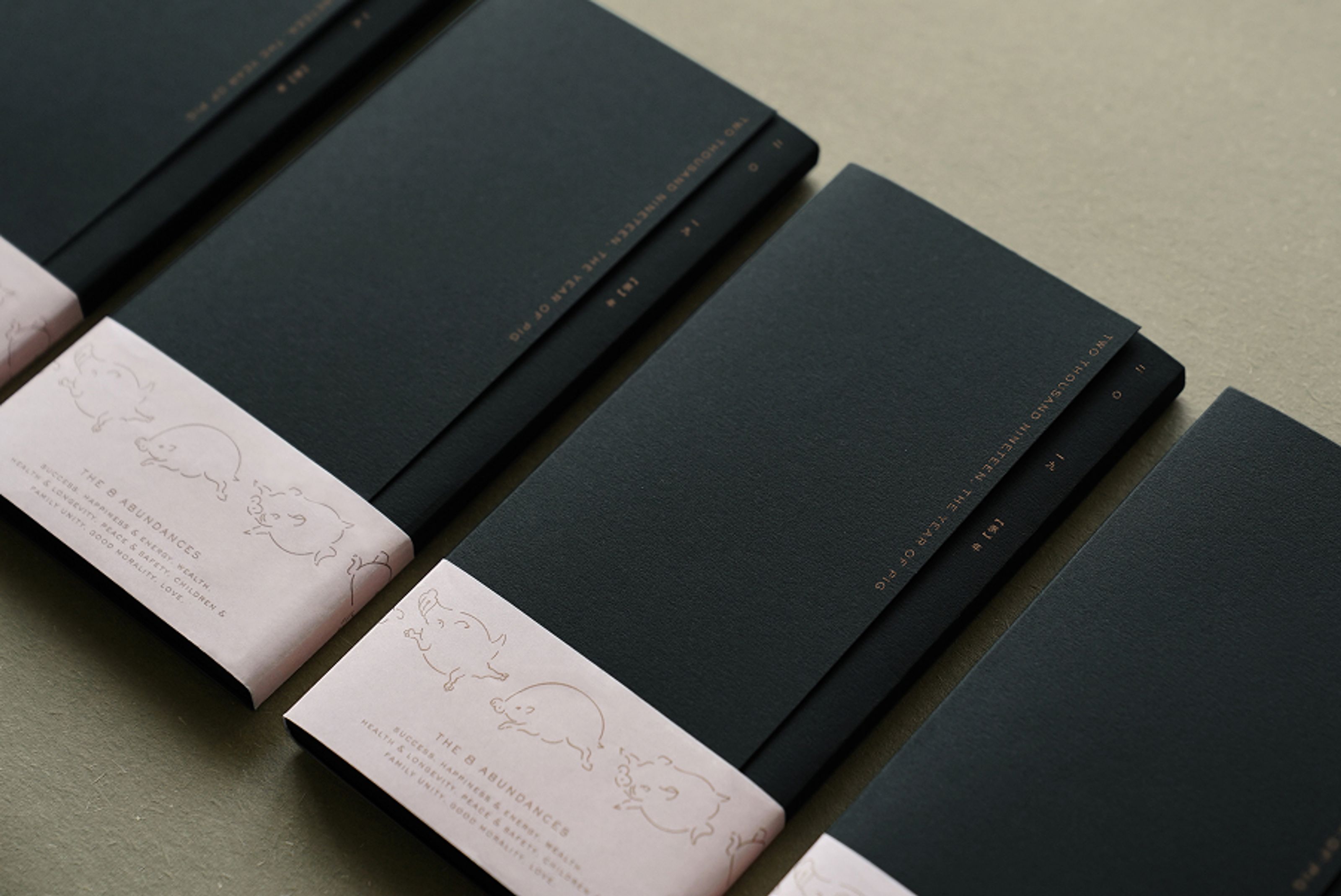 Dark gray booklets with pink bands featuring pig drawings and "The 8 Abundances" and "Year of Pig" text.