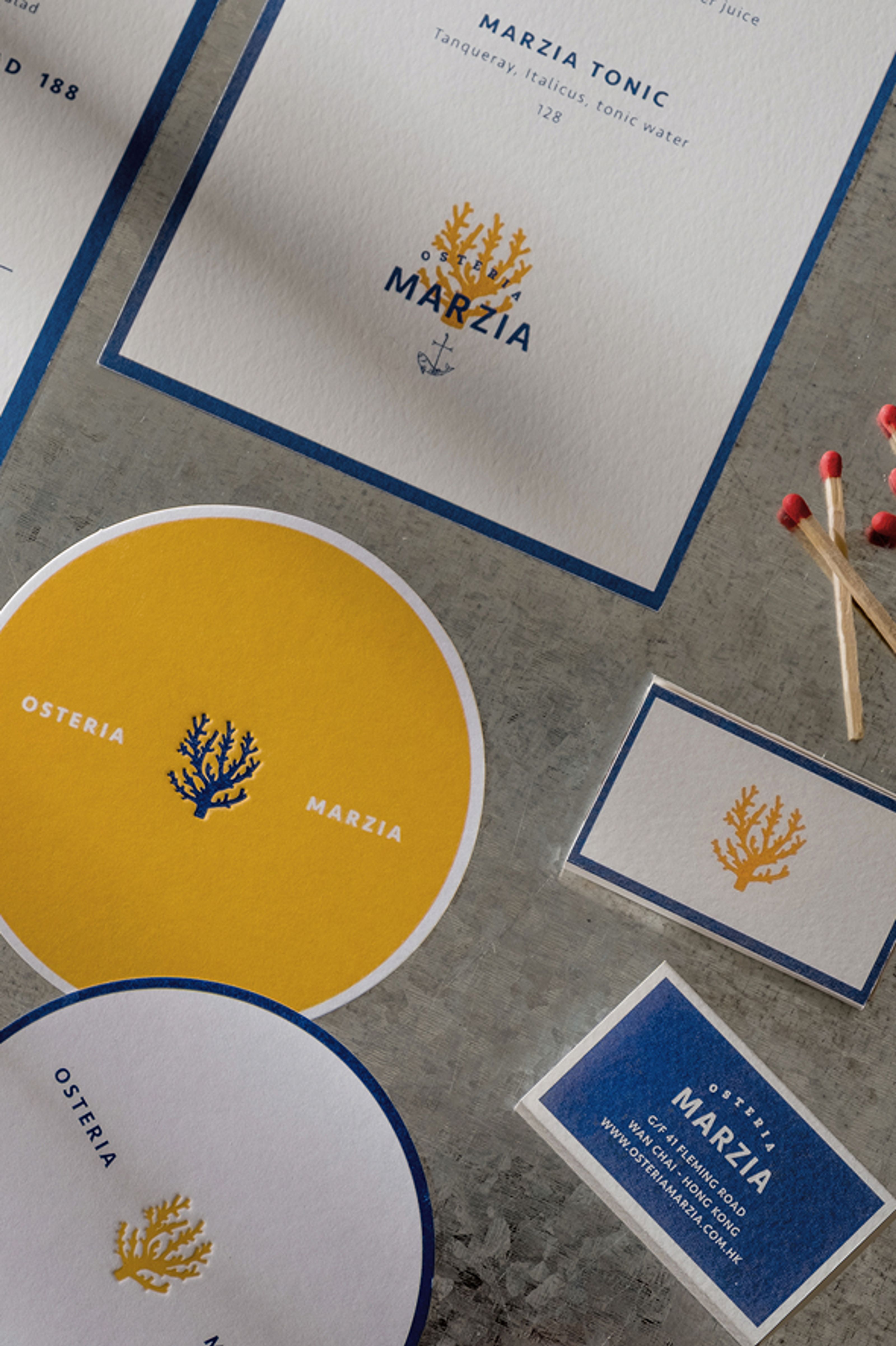 Restaurant branding materials for Osteria Marzia, including menus, coasters, and cards with coral and anchor logos in white, blue, and yellow. Red-tipped matches are also visible.