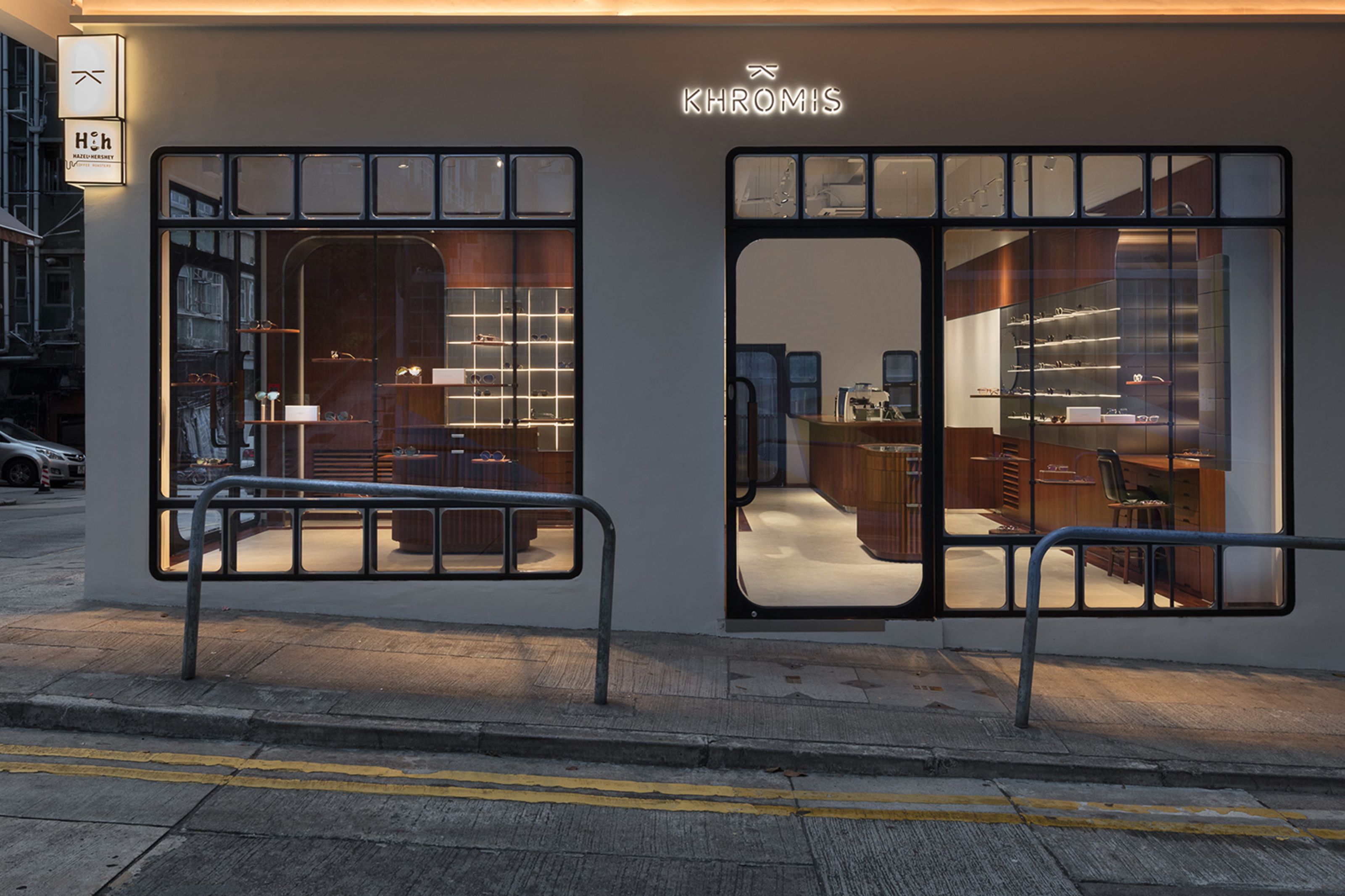 Illuminated Khromis storefront with an HcH sign, large windows displaying a warm wooden interior, and a railing on the sidewalk.