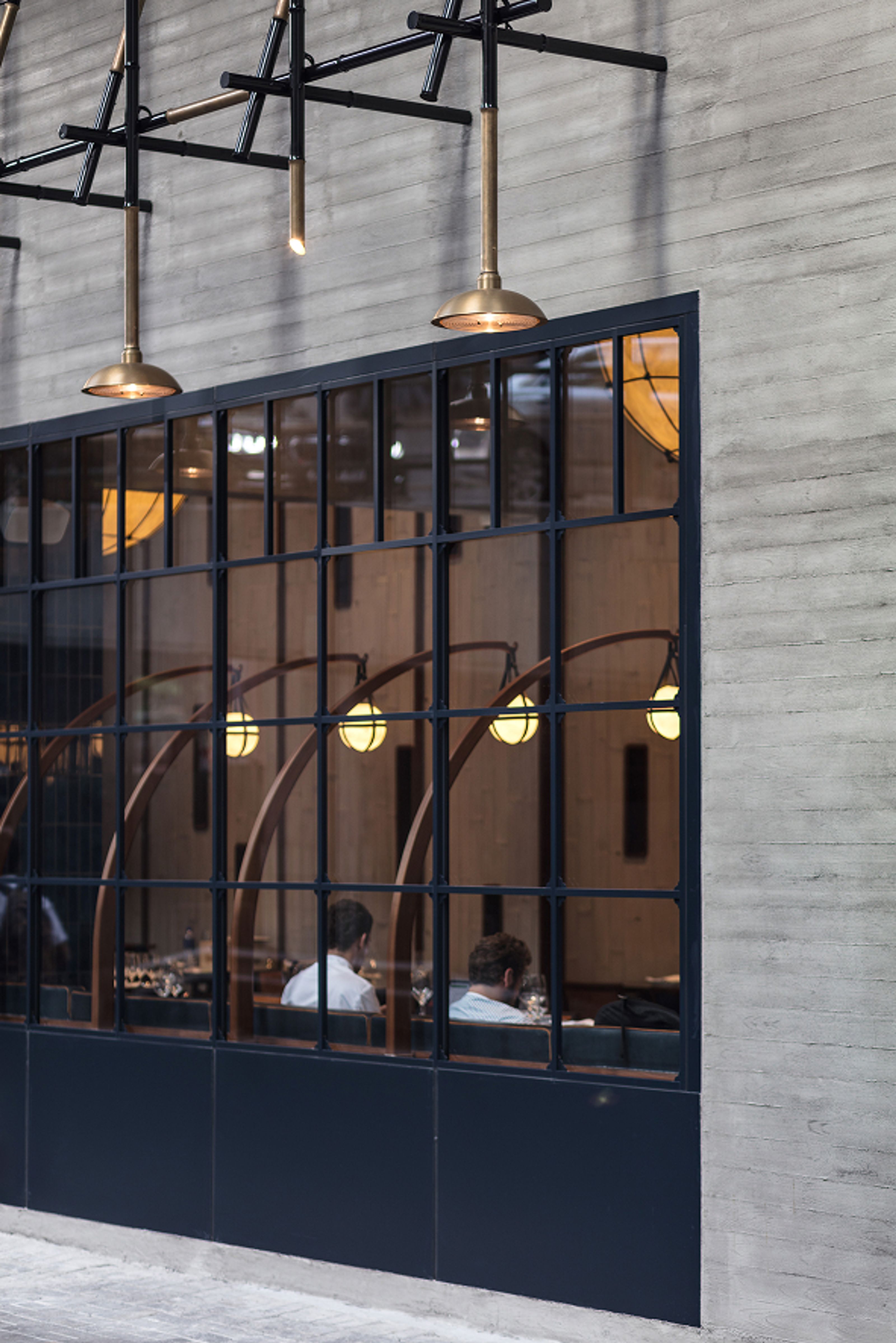 An industrial-style restaurant exterior with a grid window revealing diners and globe lights inside, framed by a concrete wall and outdoor brass lamps.