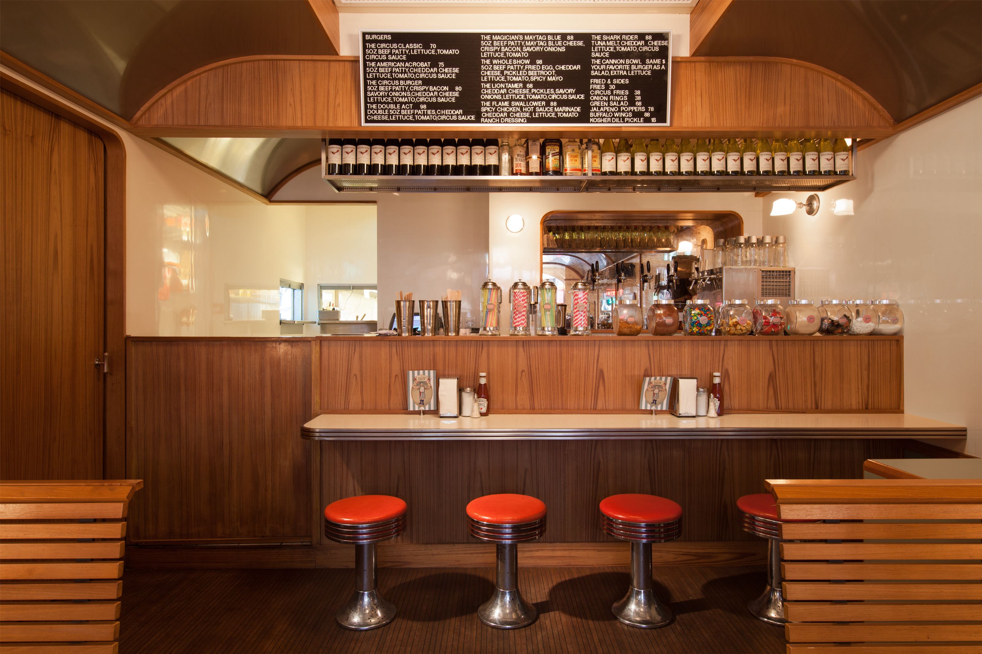 A retro diner counter area with red stools, a menu board, and jars of toppings and condiments.
