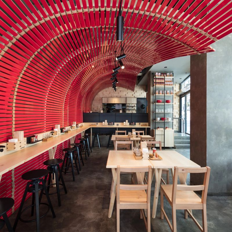 Modern restaurant interior with a curved red slatted ceiling and wall, light wood tables, and black stools.