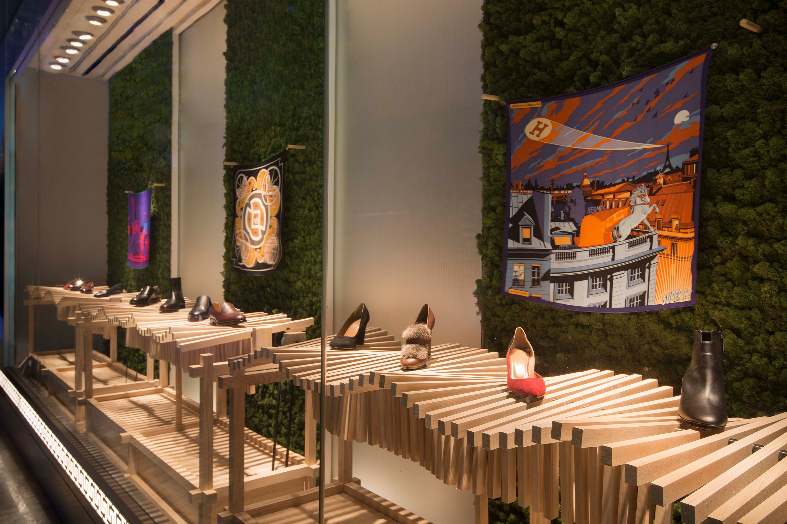 A store window display featuring shoes on elaborate wooden slat platforms, flanked by green moss walls and colorful patterned scarves.