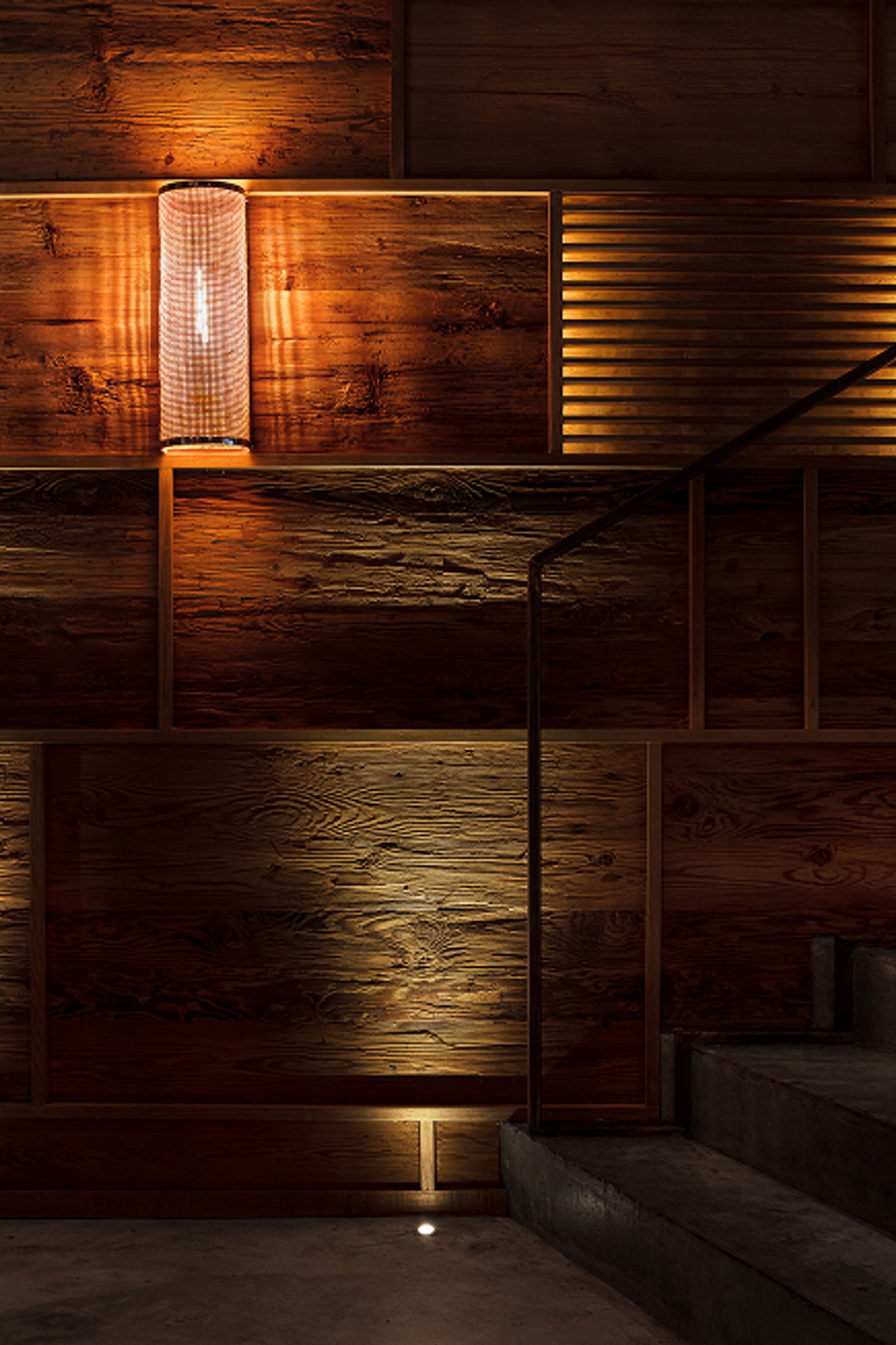 Warmly lit wooden paneling with a cylindrical lamp, accent lights, a metal handrail, and concrete steps.