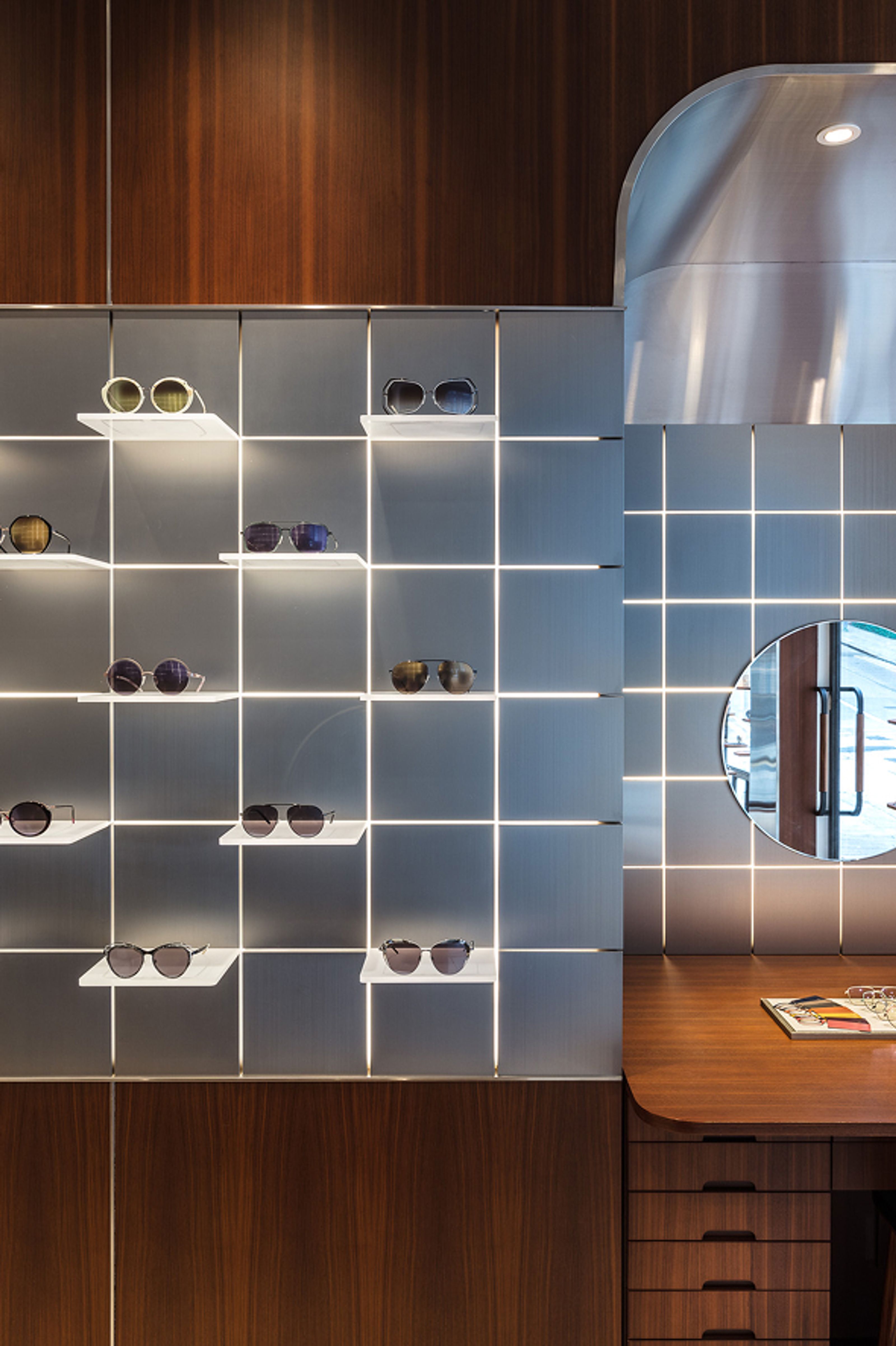 Modern optical shop interior with a grid-lit wall displaying sunglasses, a wooden desk, and a round mirror.