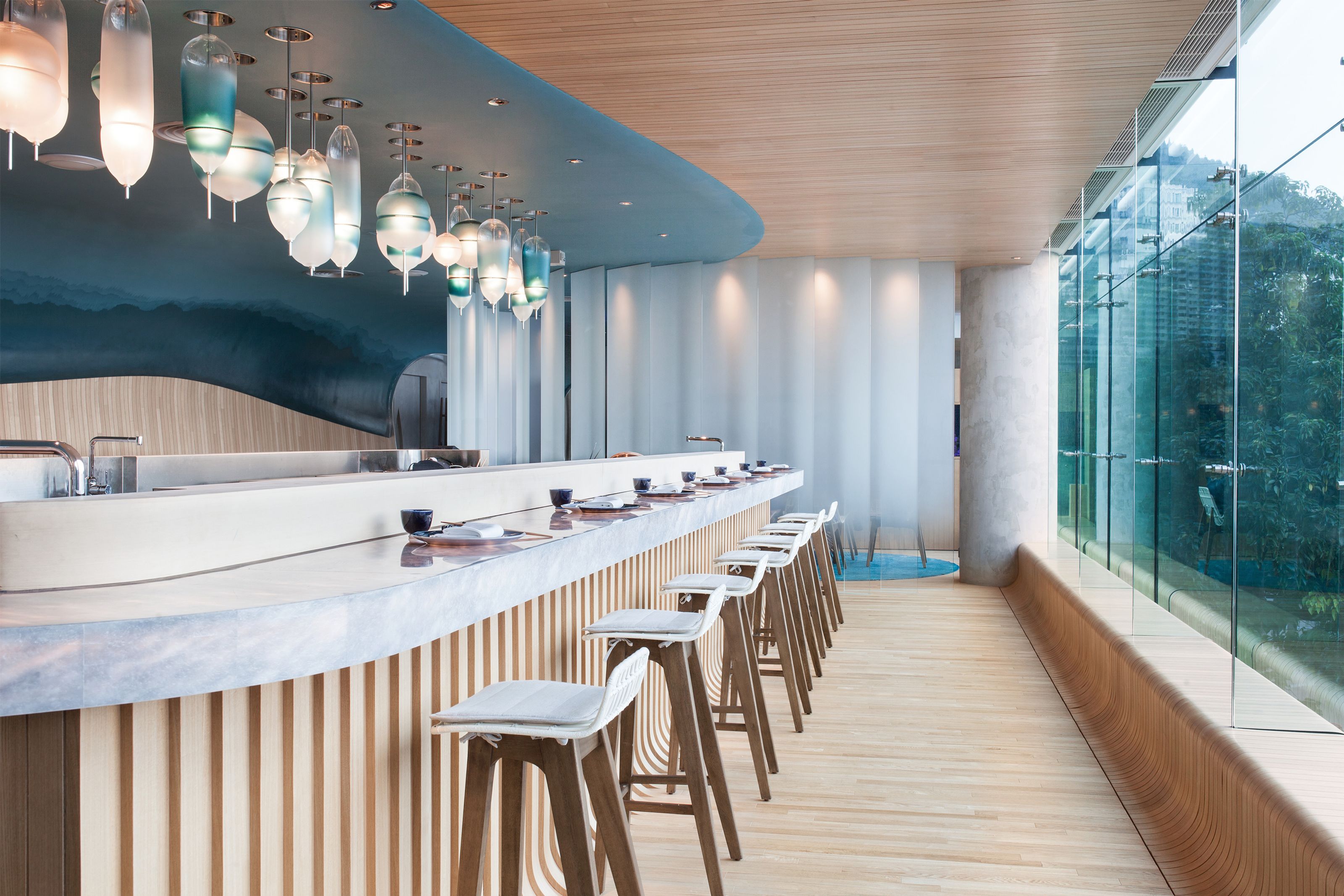 A bright, modern restaurant interior with a long wooden bar, white stools, and a blue ceiling with glass pendant lights, next to large windows overlooking trees.