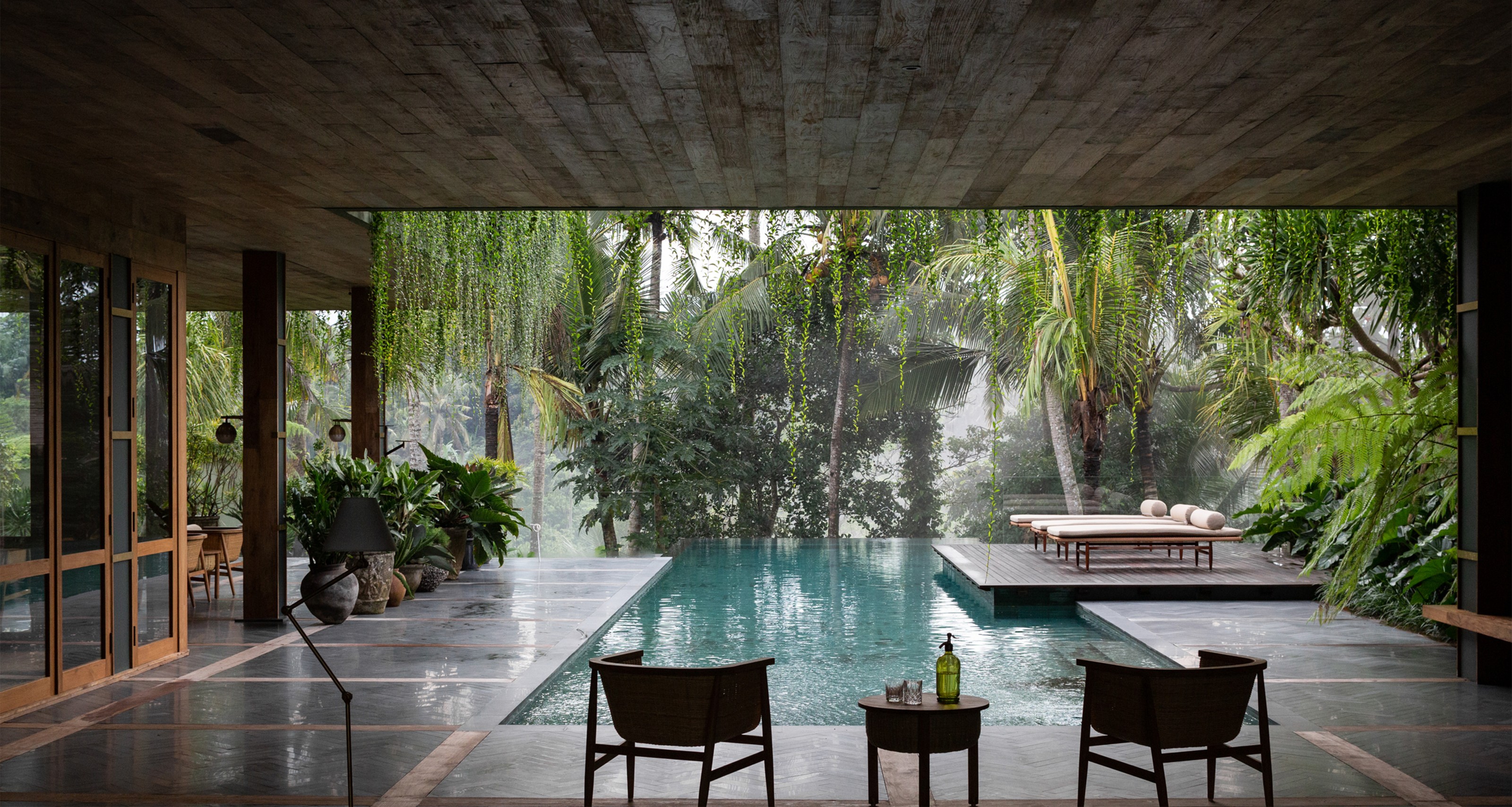 A modern, open-air concrete room overlooks a swimming pool and lush tropical jungle.