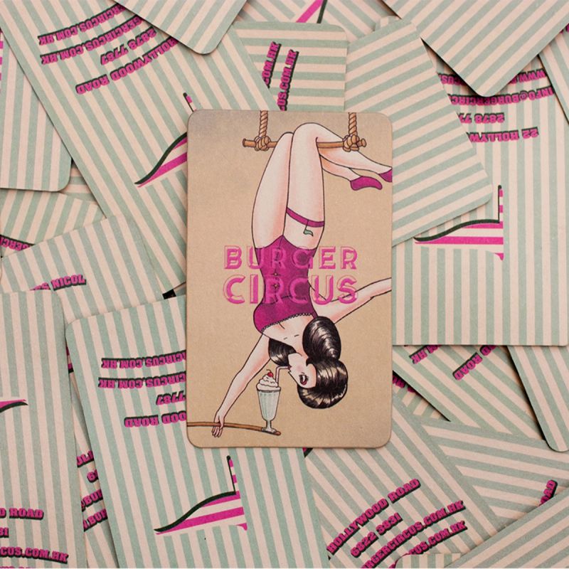 A "Burger Circus" business card featuring a pin-up style woman on a trapeze drinking a milkshake, resting on a pile of striped cards.