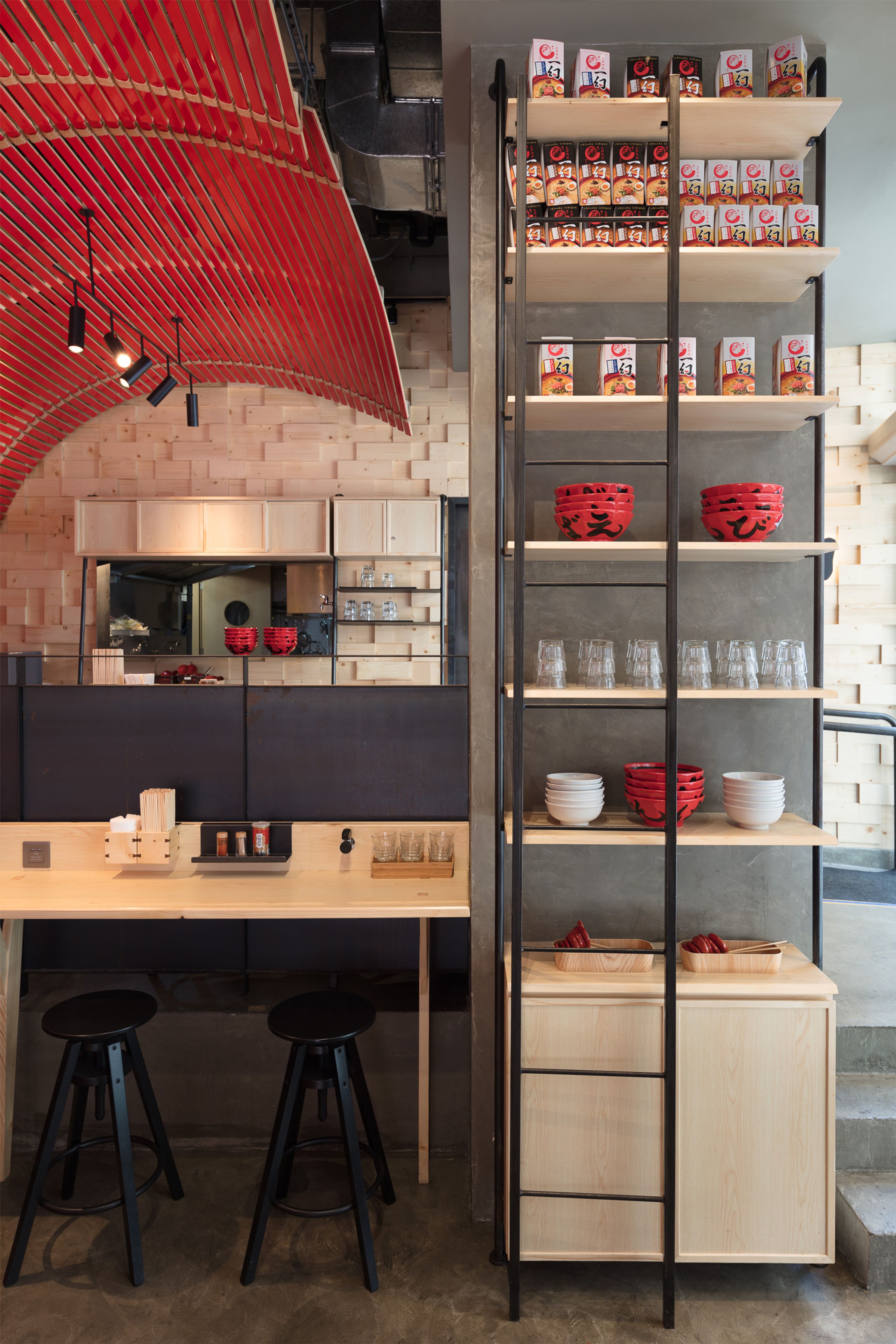 A modern ramen restaurant interior featuring a red slatted ceiling, wooden walls, a light wood counter with two black stools, and a tall shelf unit displaying ramen and bowls.