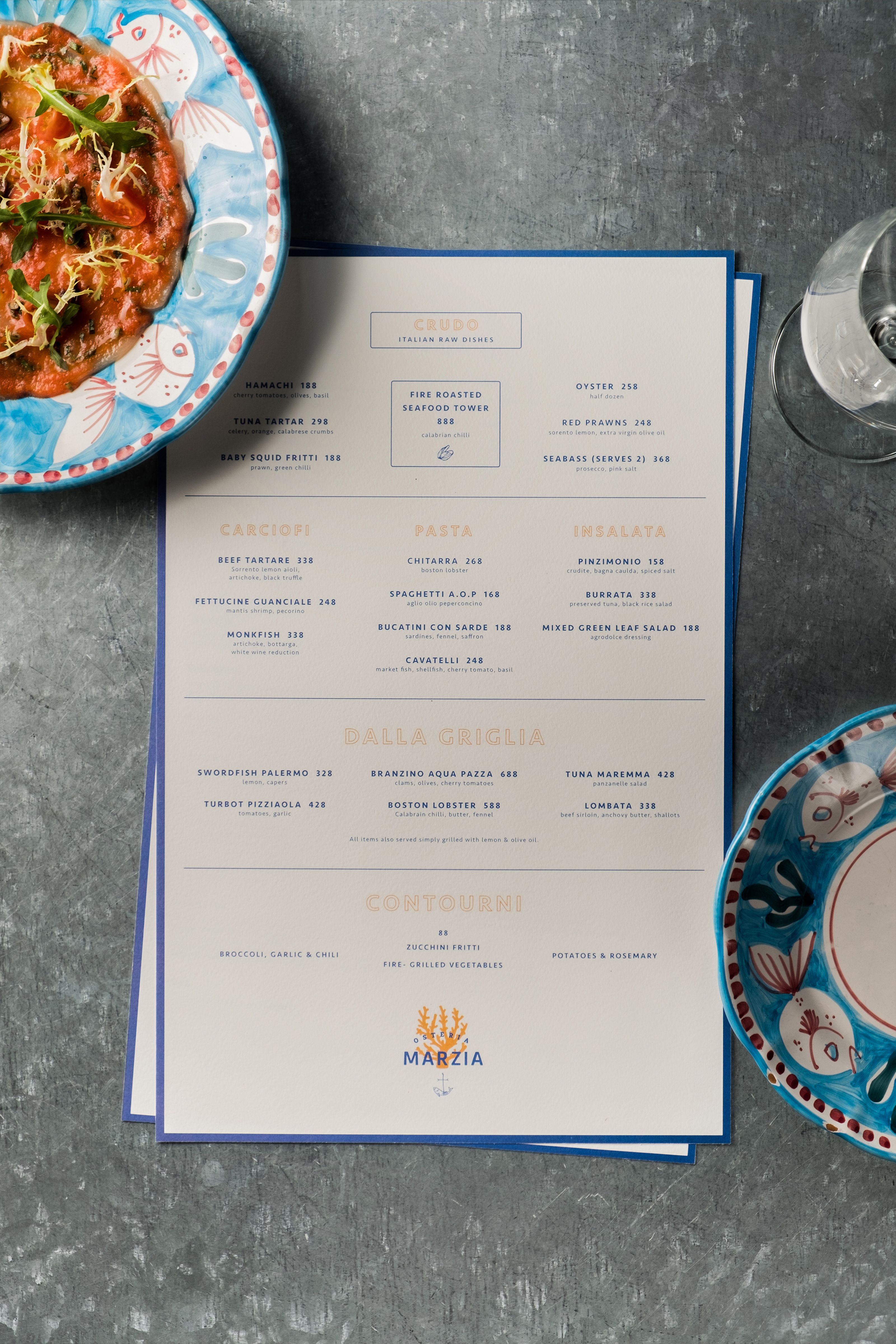 A restaurant menu for Marzia with a seafood dish, an empty wine glass, and an empty plate on a dark table.