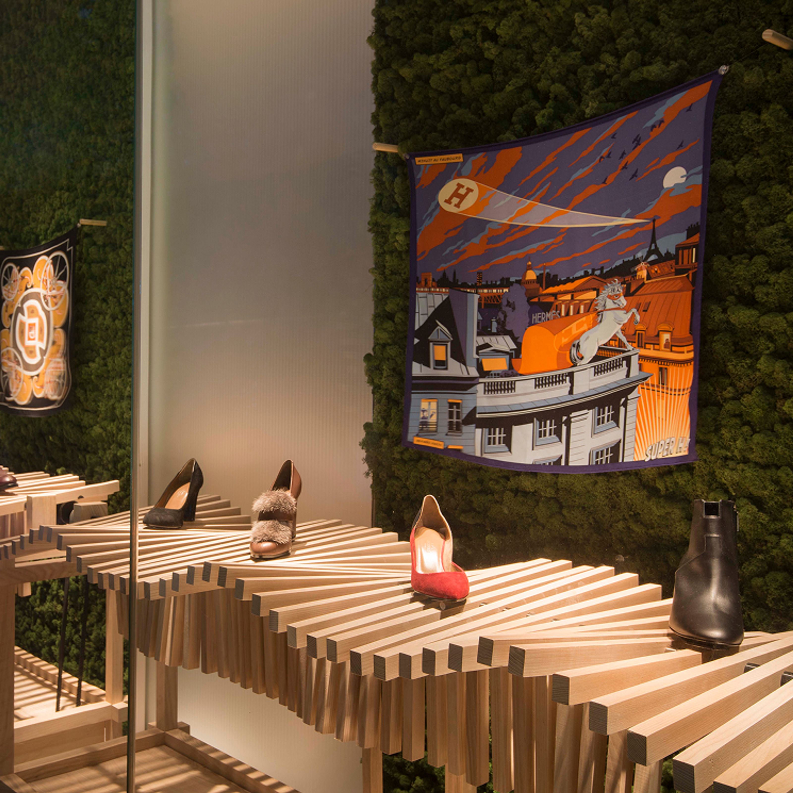 A store display with several pairs of shoes on a tiered wooden block structure against a green moss wall, featuring two decorative scarves.