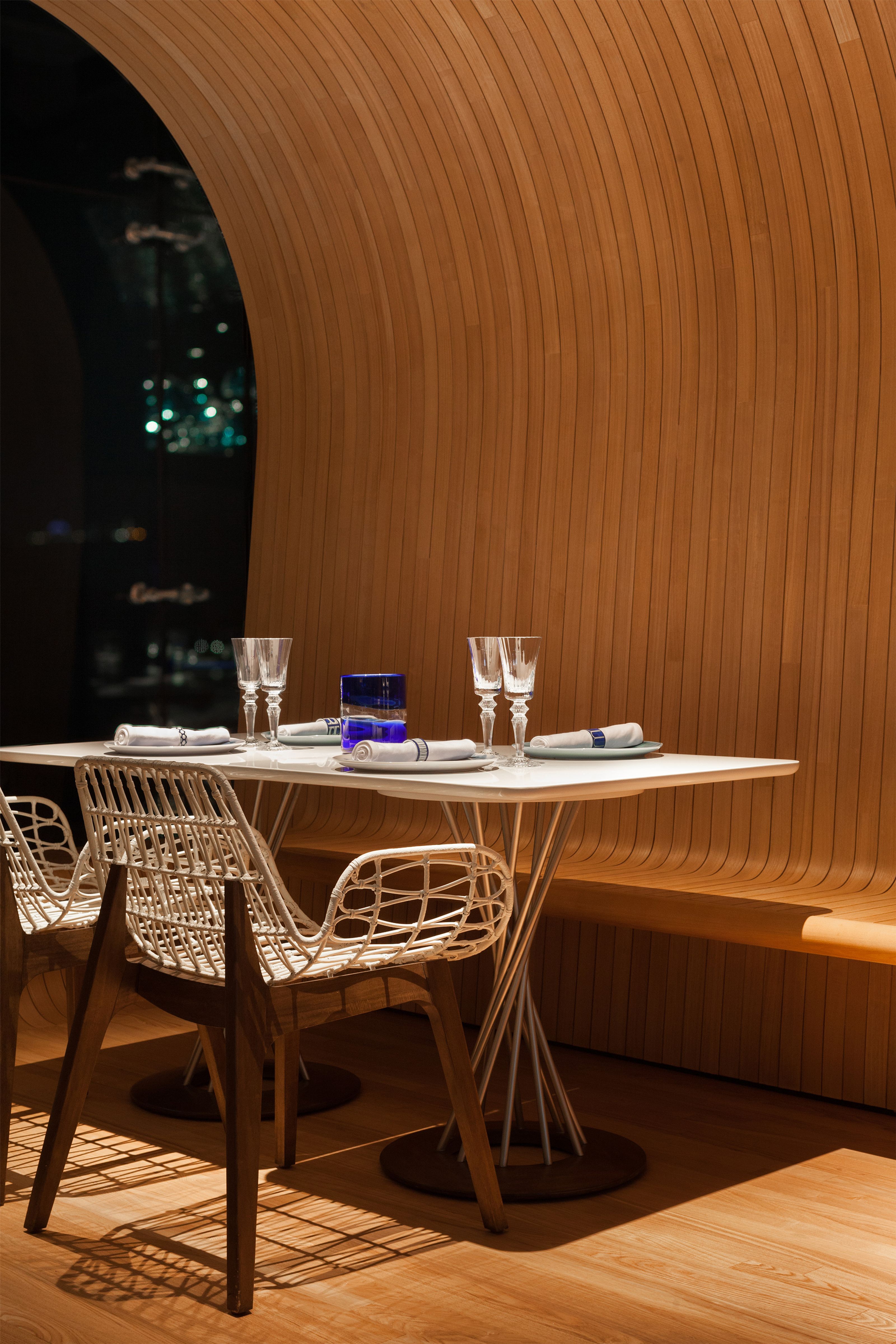 A modern restaurant table set for two with white chairs against a curved wooden wall and a dark window showing city lights.