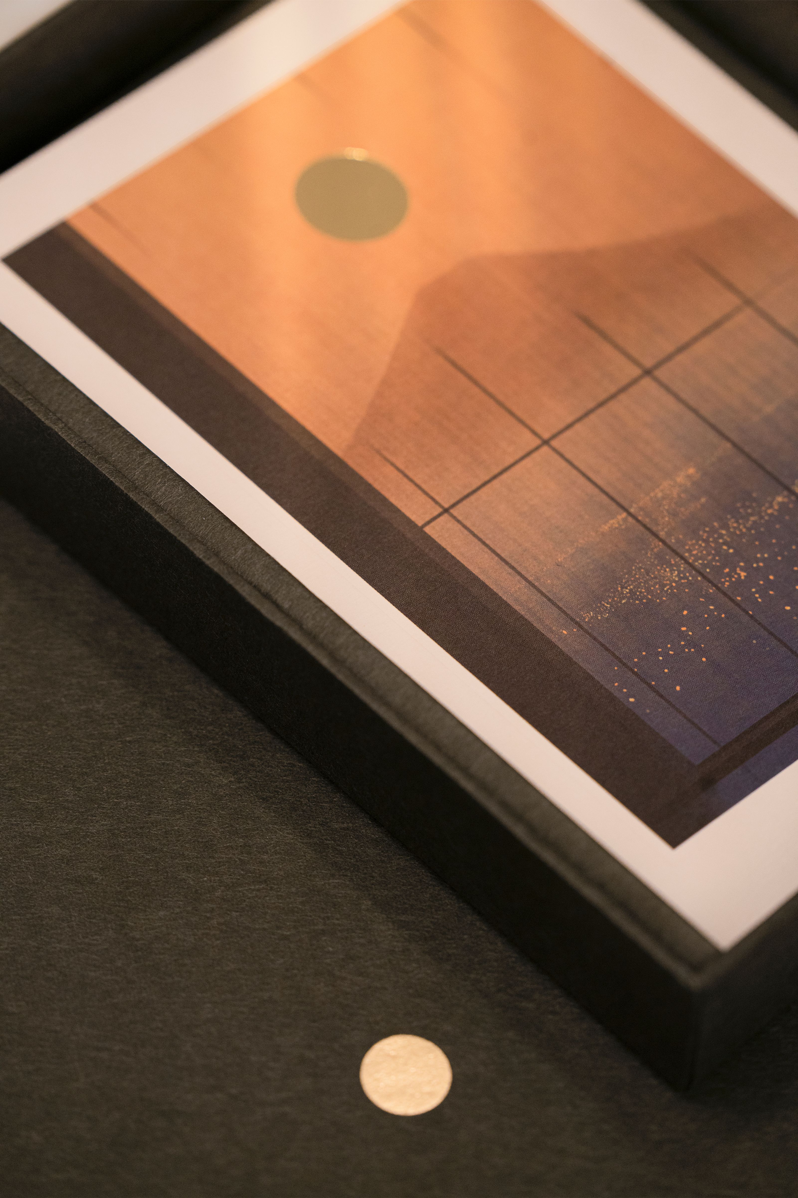 An angled view of a framed artwork with an orange to blue gradient, grid lines, and a gold foil circle, with another gold circle on a dark surface.