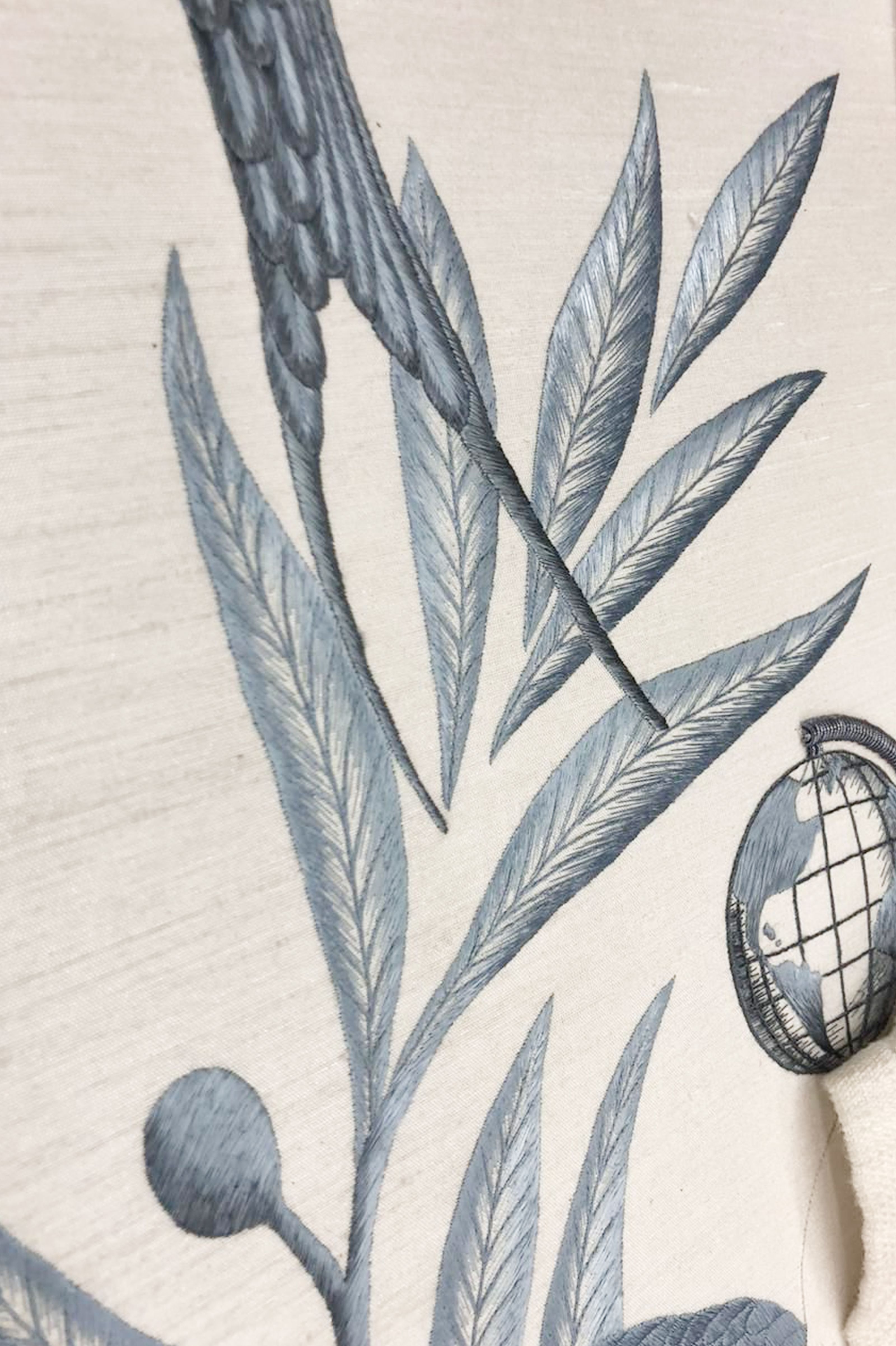 Intricate blue embroidery on white fabric, depicting leaves, a bird, and a globe.