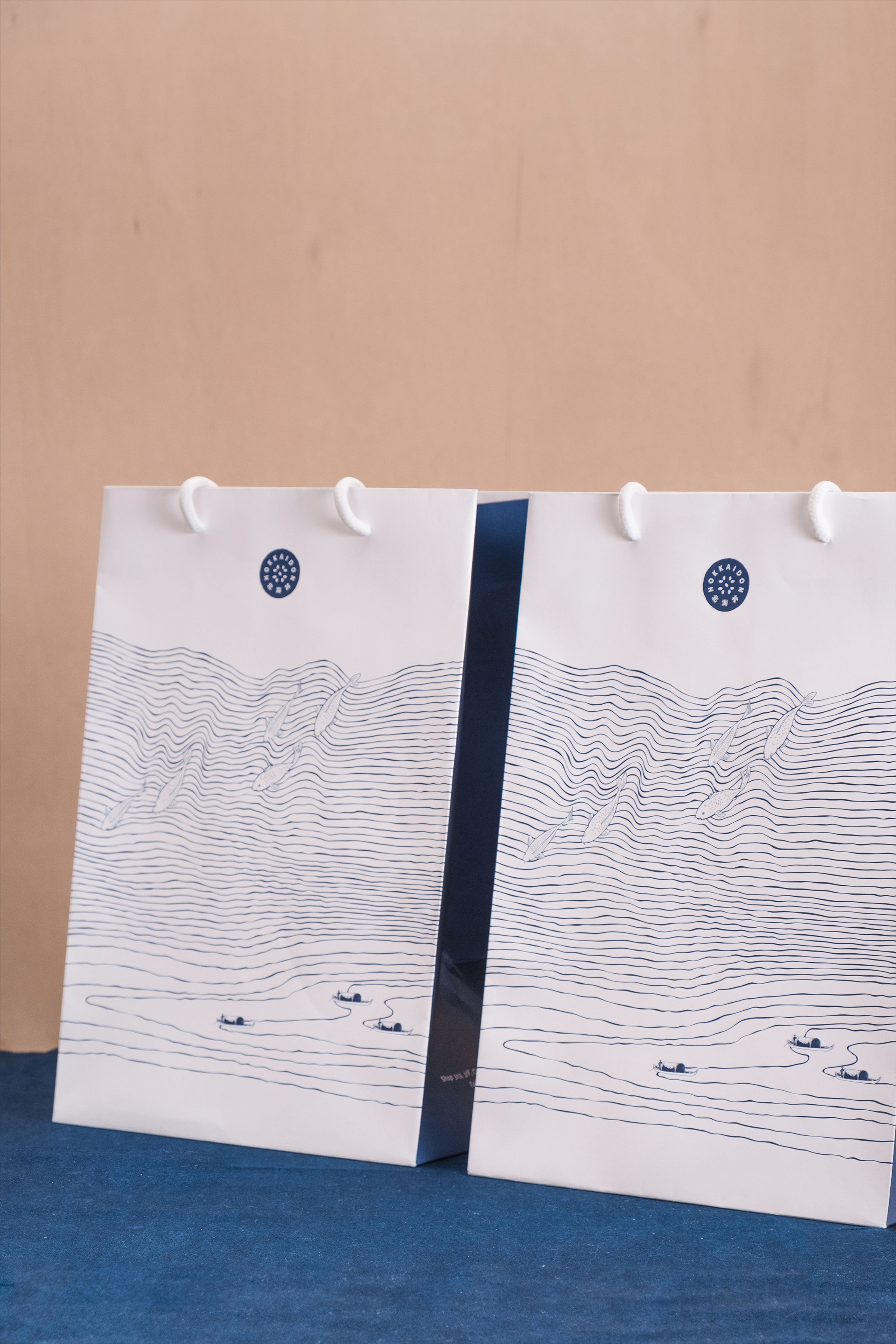 Two white paper bags with blue line art depicting waves, fish, and small boats, and a circular blue logo.