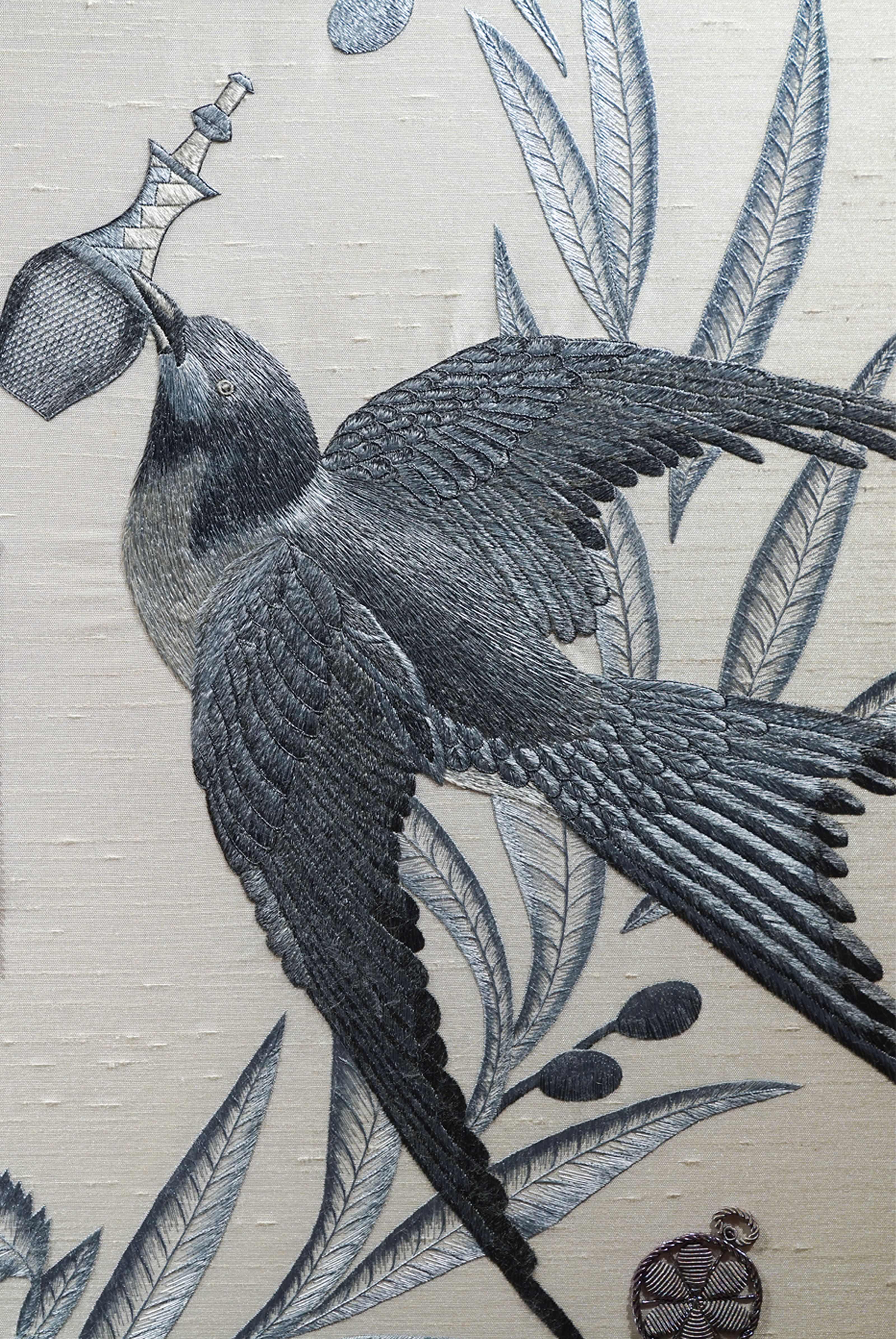 Detailed grey embroidery of a bird flying upwards with an ornate silver bottle in its beak, on a light fabric background with leaves.