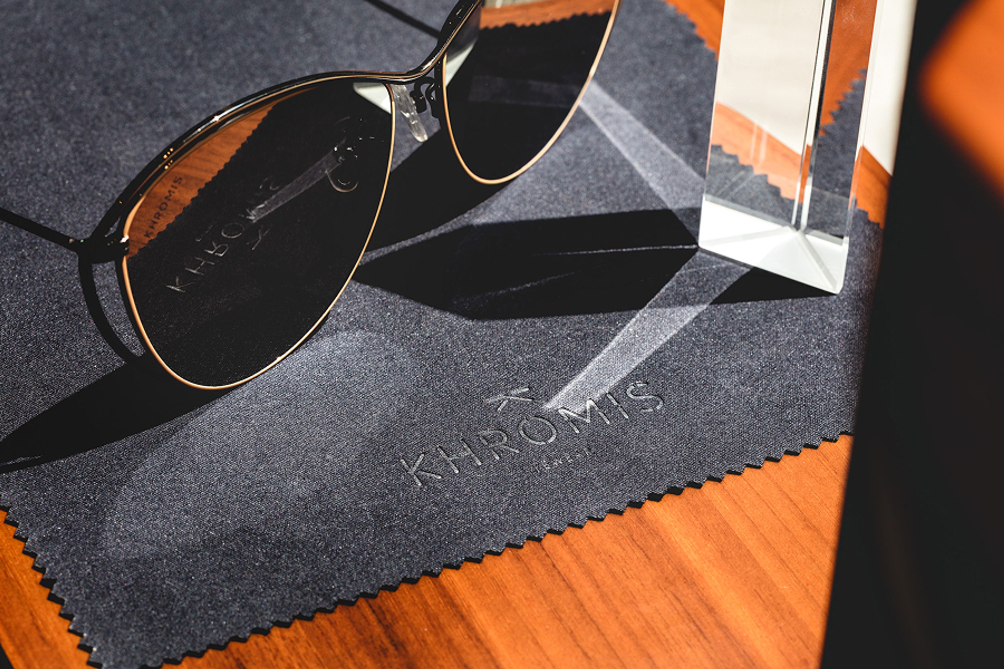 Gold-rimmed sunglasses rest on a dark grey cleaning cloth with a "KHROMIS" logo, next to a prism creating light patterns on a wooden table.