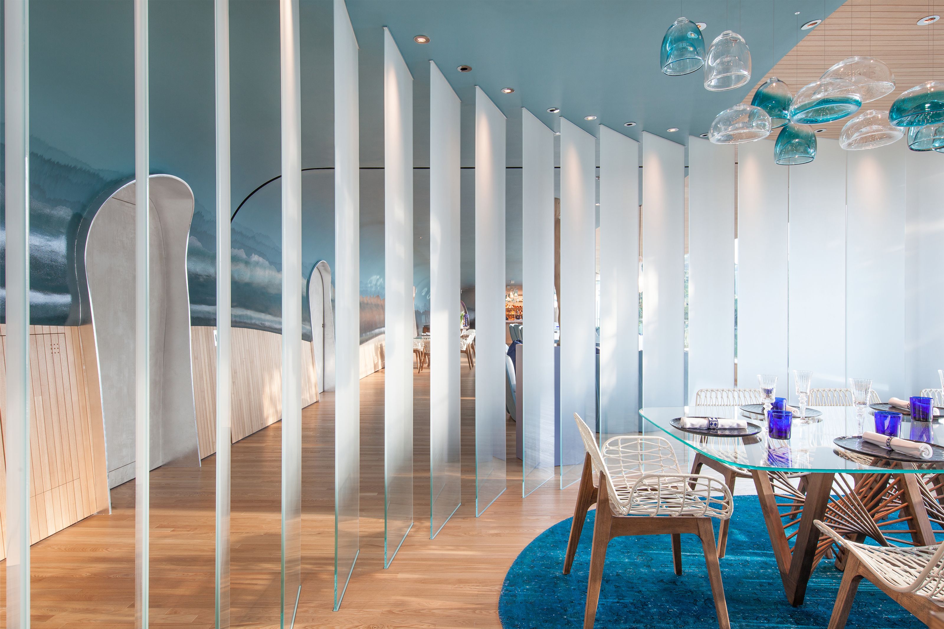 Modern restaurant interior with a glass dining table, translucent dividers, and a wavy blue accent wall with suspended glass lights.