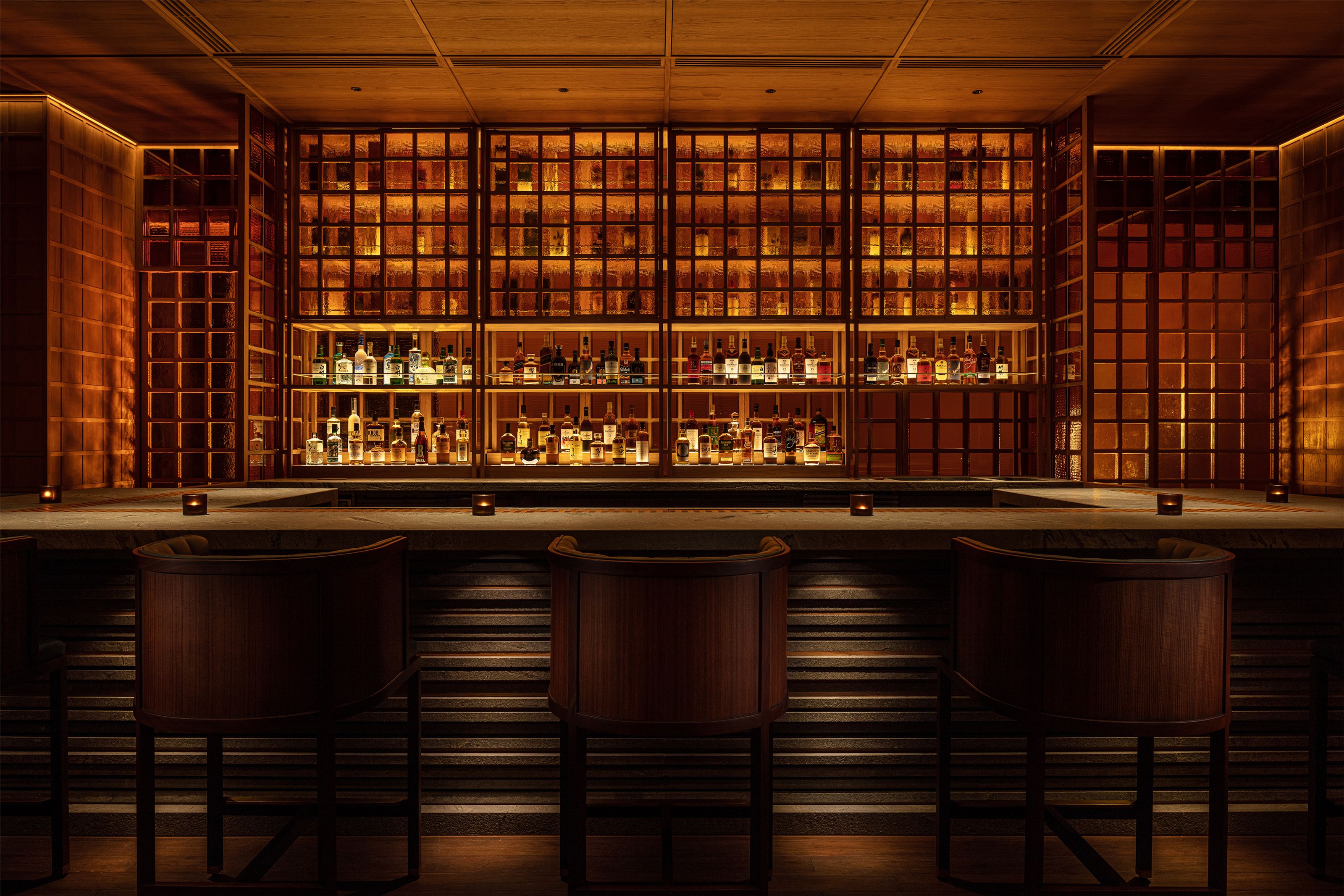 A warm, dimly lit bar with three stools facing a large, backlit shelving unit filled with liquor bottles.